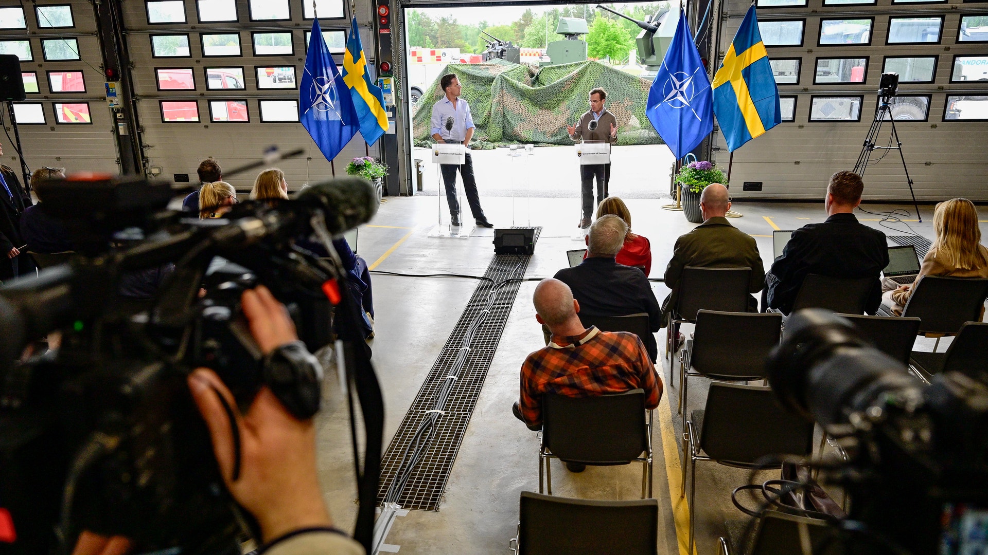 En presskonferens eller offentligt evenemang med åskådare som sitter och tittar på två personer som står på en scen med svenska flaggor i bakgrunden.
