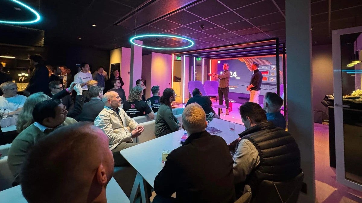 En grupp människor minglar på ett event i ett modernt rum med blå och lila belysning. Flera personer står vid höga bord och diskuterar medan andra rör sig omkring i rummet.