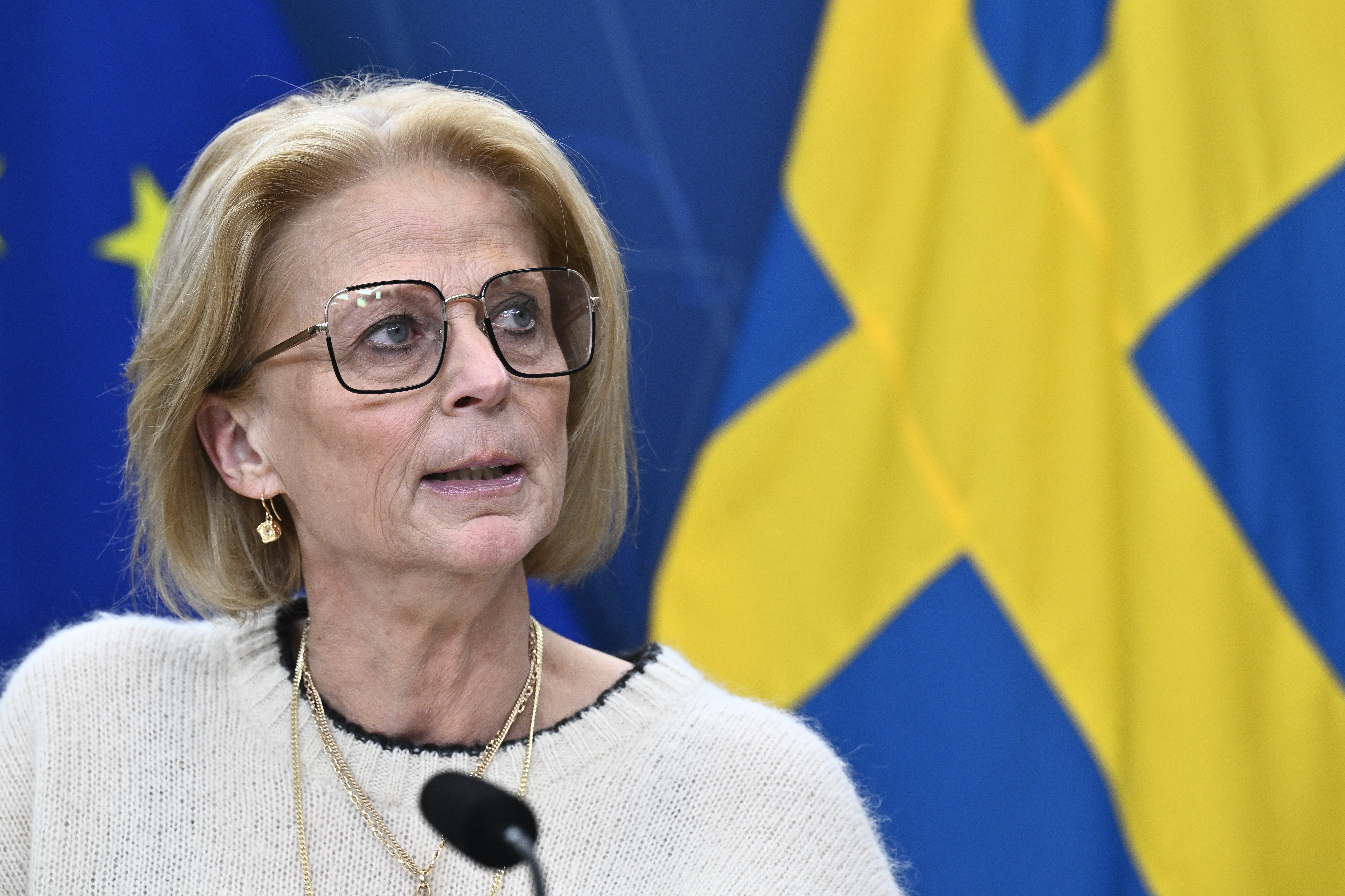 Elisabeth Svantesson med glasögon och ljus tröja talar i en mikrofon framför en svensk flagga och en EU-flagga.