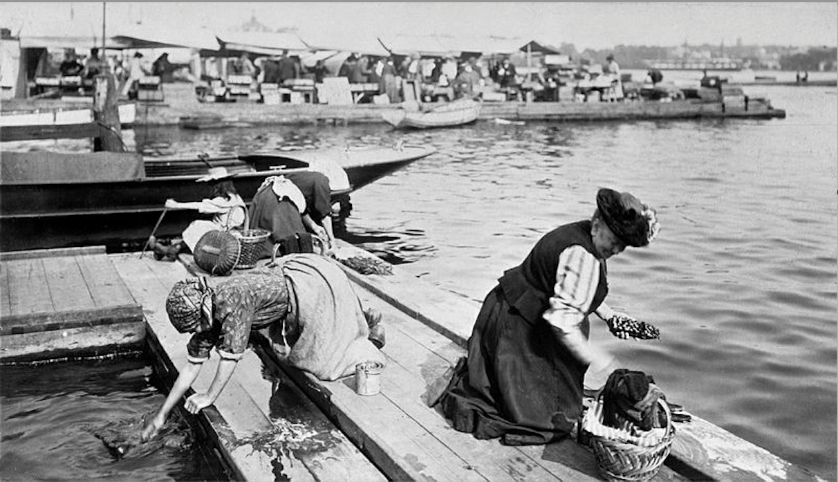 Svartvit fotografi från en hamn där människor arbetar på en båt vid kaj.