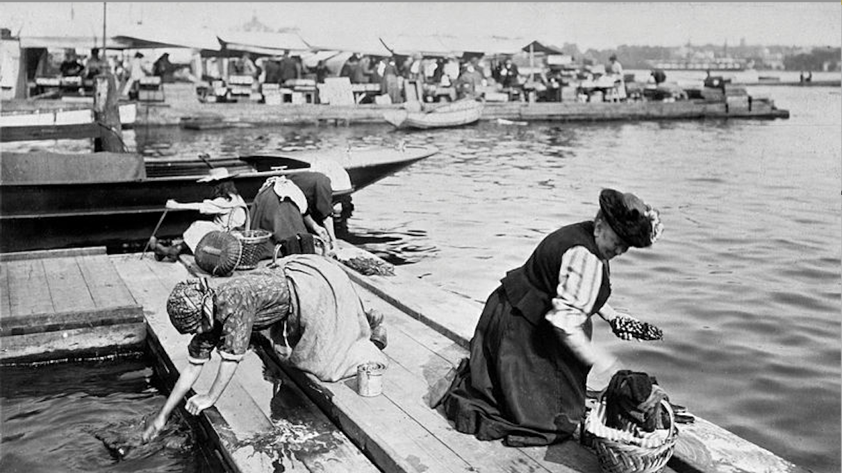 Svartvit fotografi från en hamn där människor arbetar på en båt vid kaj.
