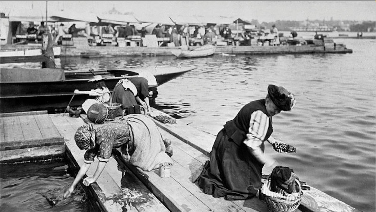 Svartvit fotografi från en hamn där människor arbetar på en båt vid kaj.