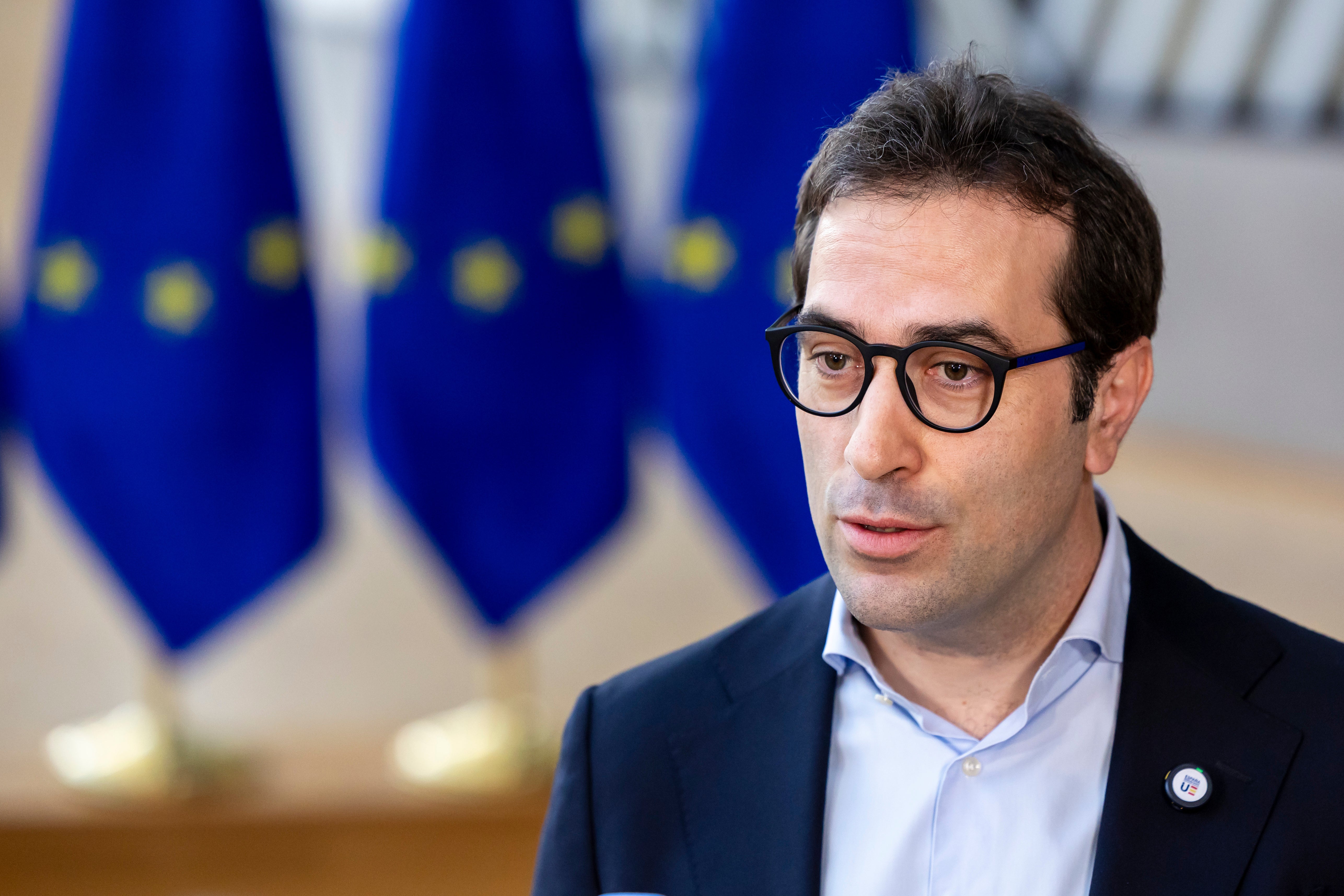 Porträttfoto av en man med glasögon i mörkblå kavaj framför EU-flaggor