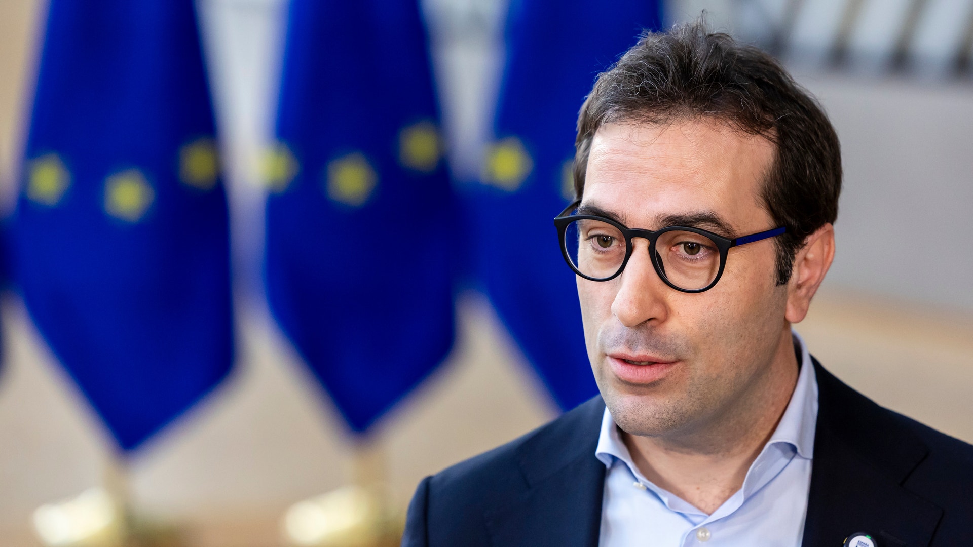 Porträttfoto av en man med glasögon i mörkblå kavaj framför EU-flaggor