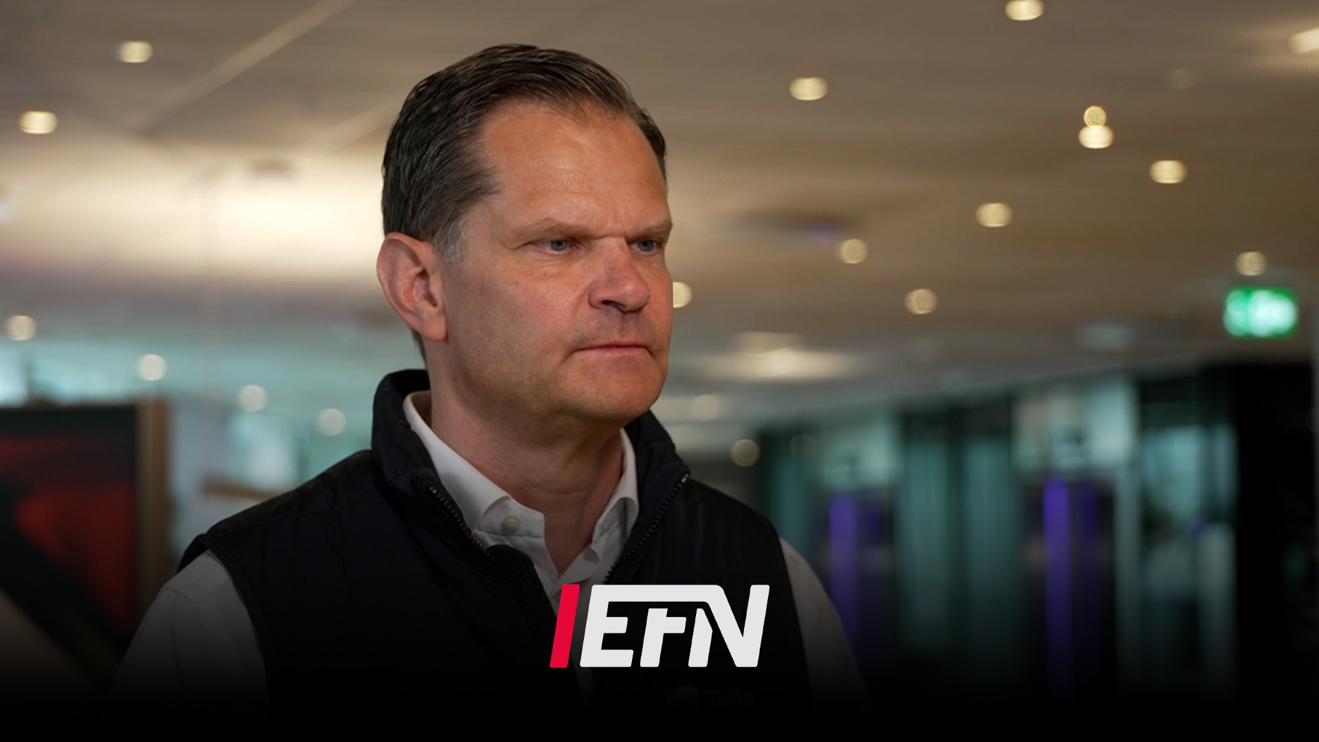 En man i svart väst står i en modern inomhusmiljö och tittar åt sidan.