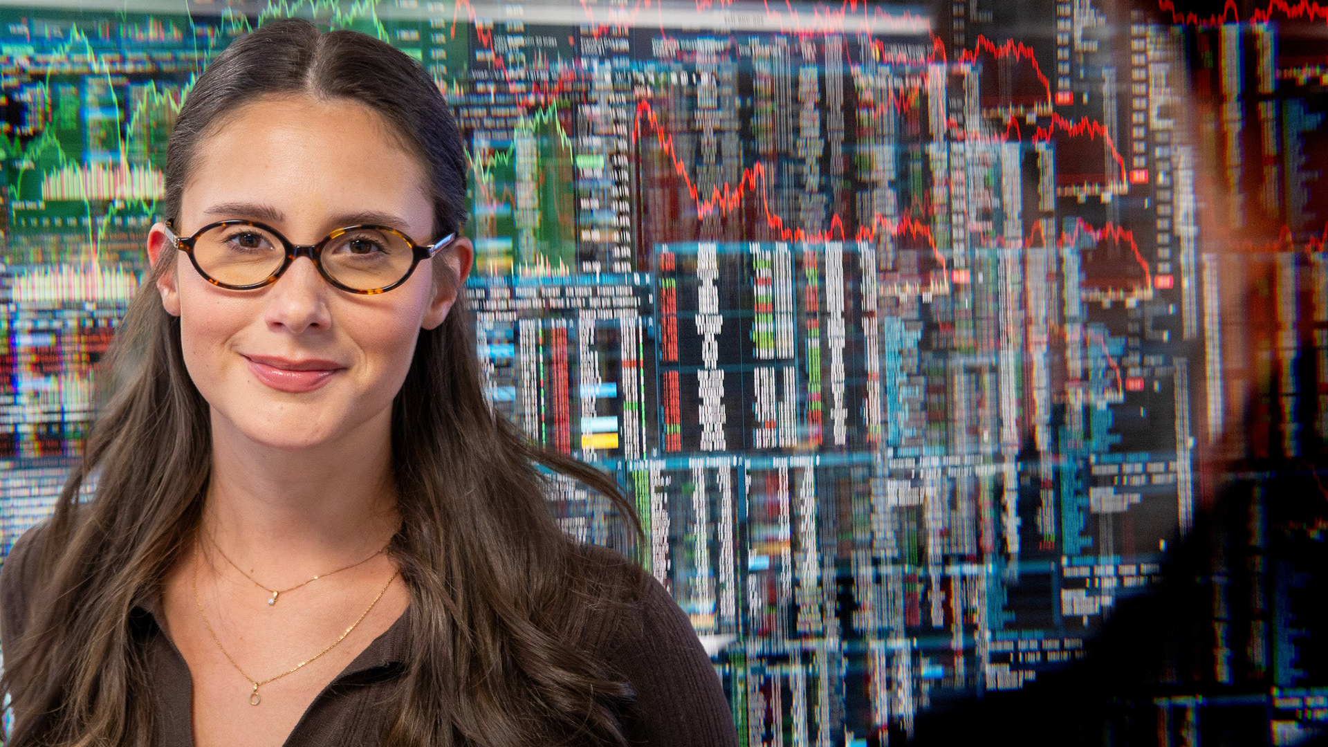 Porträtt av en kvinna med glasögon framför en bakgrund med finansiella diagram och aktiekursgrafer.