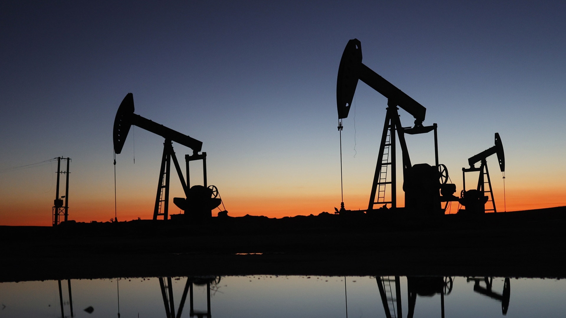 Silhuetter av oljepumpar mot solnedgångshimmel med reflektioner i vatten