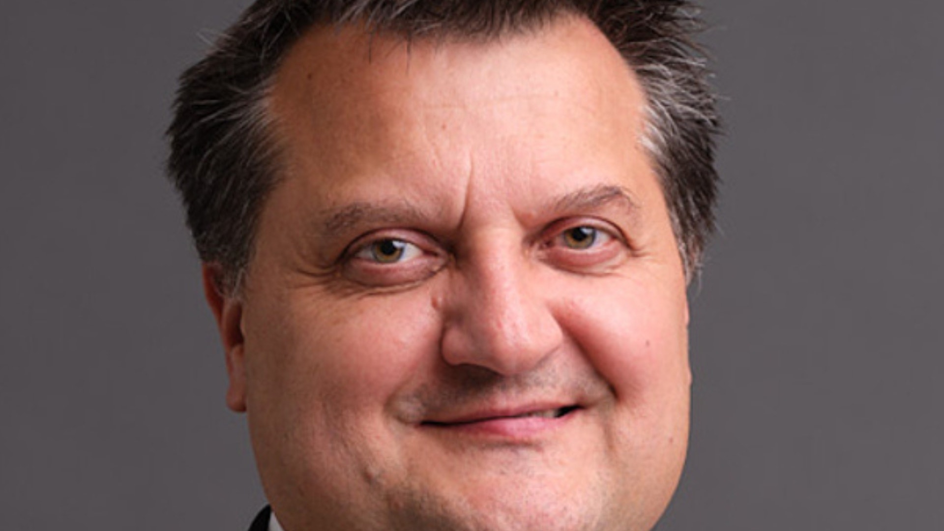 Porträtt av en man i mörk kavaj och vit skjorta mot grå bakgrund