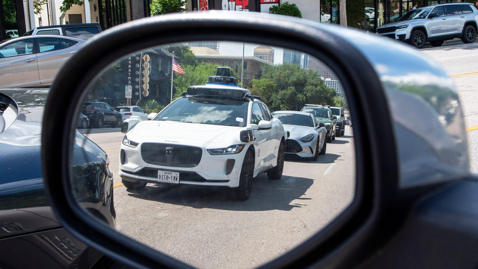 Spegelbild från en bil som visar flera vita autonoma fordon på en stadsgata. Fordonet i fokus är en Jaguar I-PACE med sensorer monterade på taket för självkörande teknik. Flera andra autonoma bilar syns i bakgrunden på en solig stadsgata med byggnader och träd.