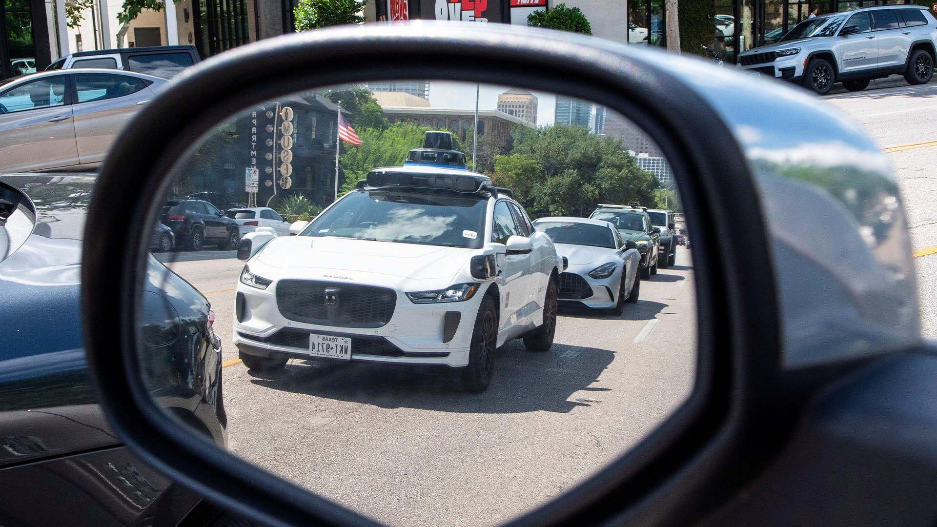 Spegelbild från en bil som visar flera vita autonoma fordon på en stadsgata. Fordonet i fokus är en Jaguar I-PACE med sensorer monterade på taket för självkörande teknik. Flera andra autonoma bilar syns i bakgrunden på en solig stadsgata med byggnader och träd.