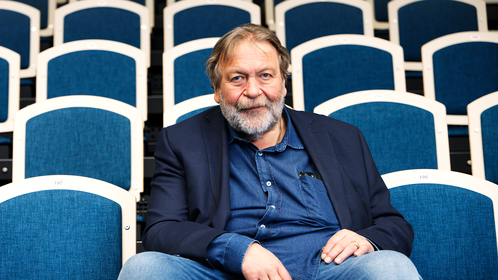 En äldre man med grått skägg sitter på en blå teaterstol. Han är klädd i mörkblå kavaj och ljusblå skjorta, och tittar mot kameran. Bakom honom syns fler tomma blå teaterstolar.
