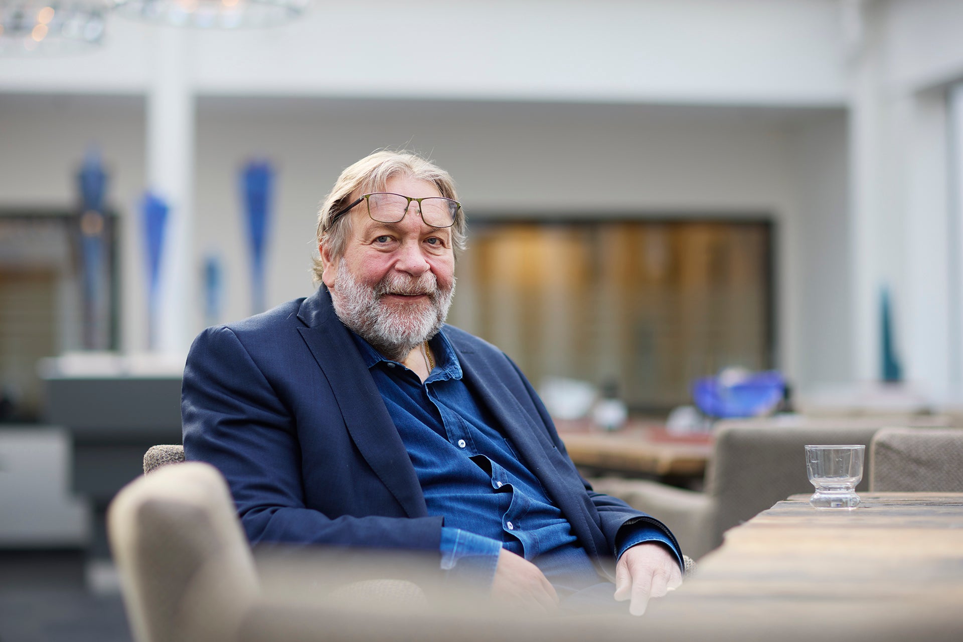 Porträtt av en äldre man med grått skägg och glasögon, klädd i mörkblå kavaj och blå skjorta, sittande i ett modernt kontor.