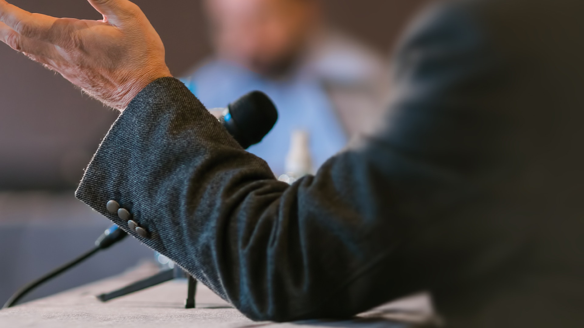 En person gestikulerar med handen framför en mikrofon under ett tal eller en intervju.