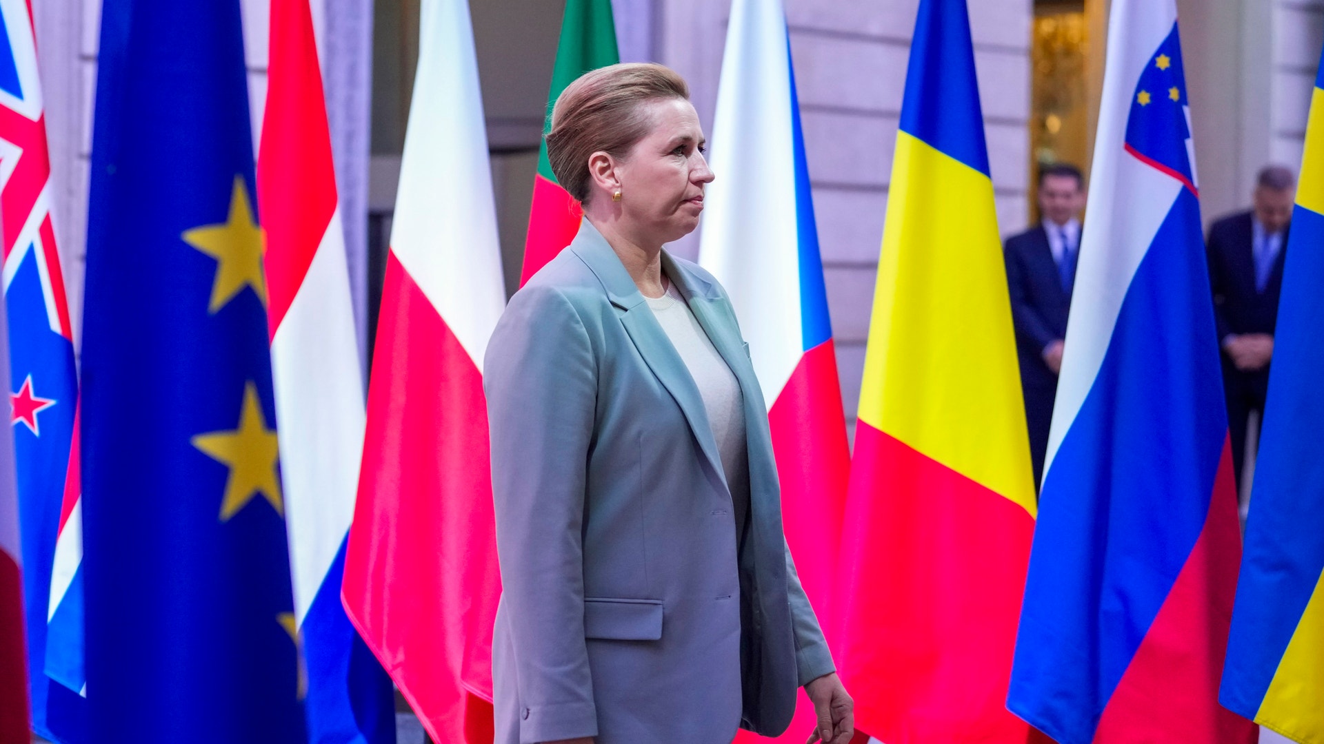 Kvinna i grå kavaj står framför internationella flaggor i en officiell miljö.