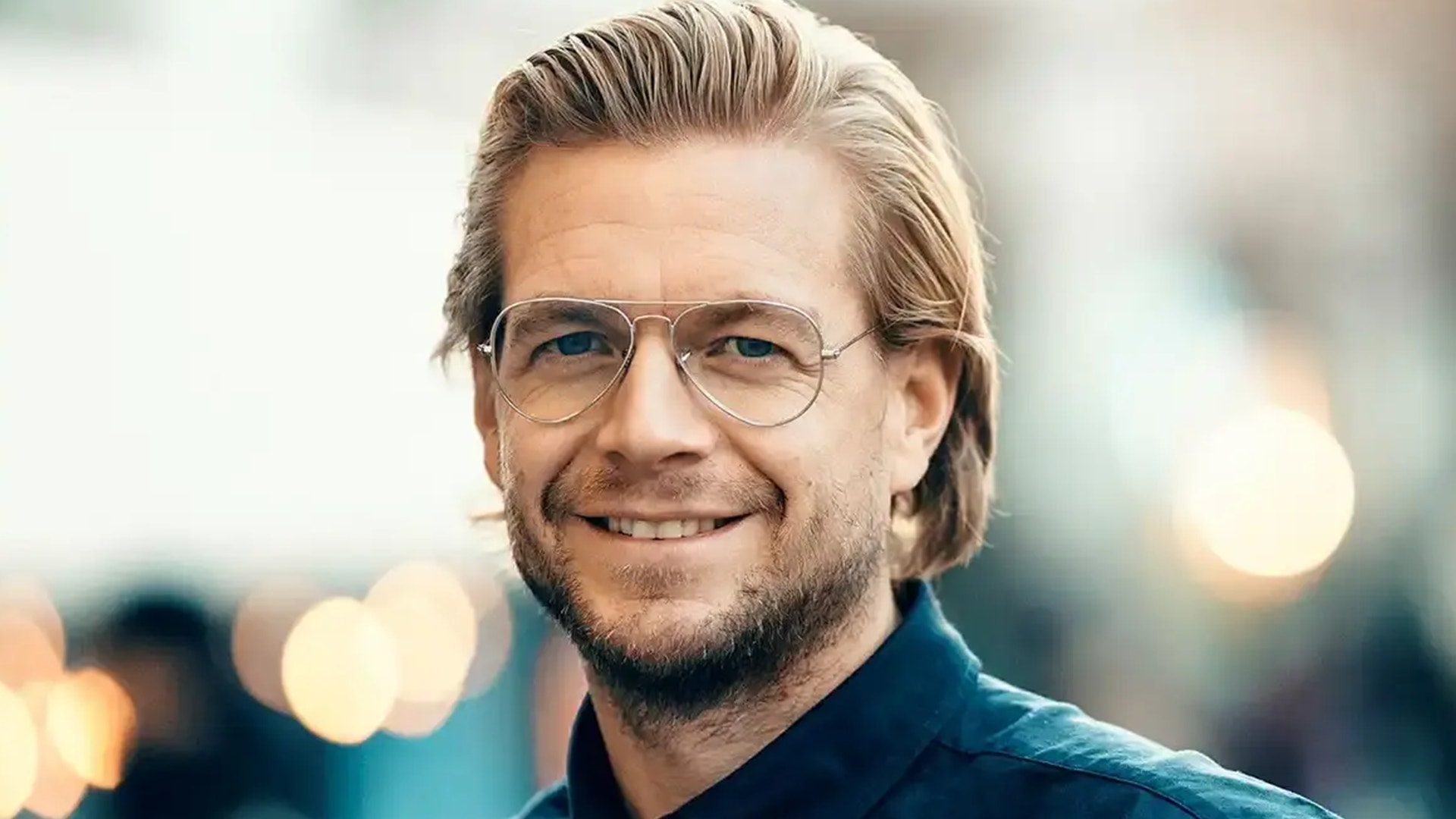 Porträtt av en Conny Ryk med glasögon och ljust hår som ler mot kameran. Bakgrunden är suddig med varma ljuseffekter.