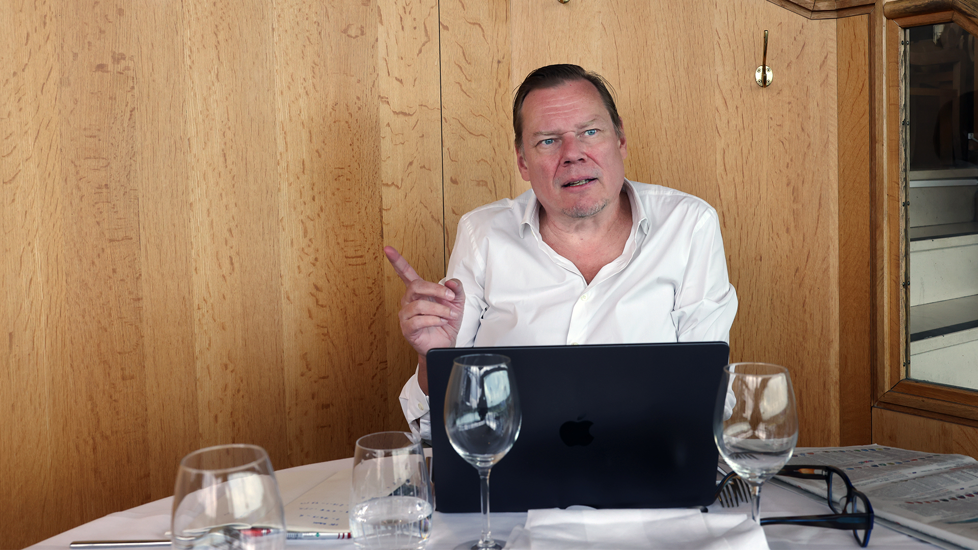Man i vit skjorta sitter vid ett bord med en laptop och presenterar eller deltar i en videokonferens.