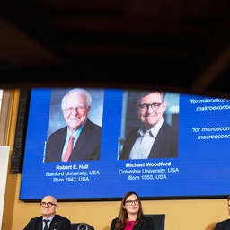 Tre personer sitter under en stor skärm som visar porträtt av två män, Robert E. Hall och Michael Woodford, vid tillkännagivandet av Nobelpriset i ekonomi.