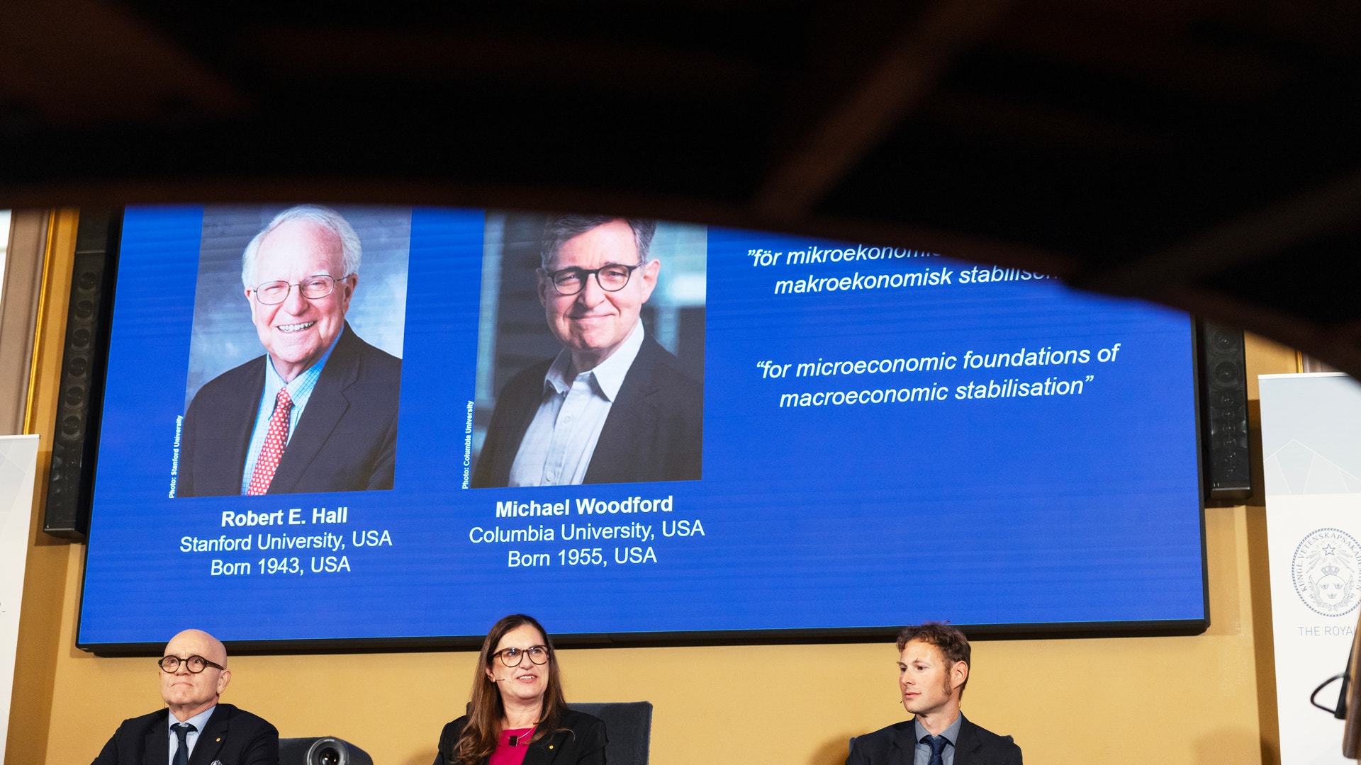 Tre personer sitter under en stor skärm som visar porträtt av två män, Robert E. Hall och Michael Woodford, vid tillkännagivandet av Nobelpriset i ekonomi.
