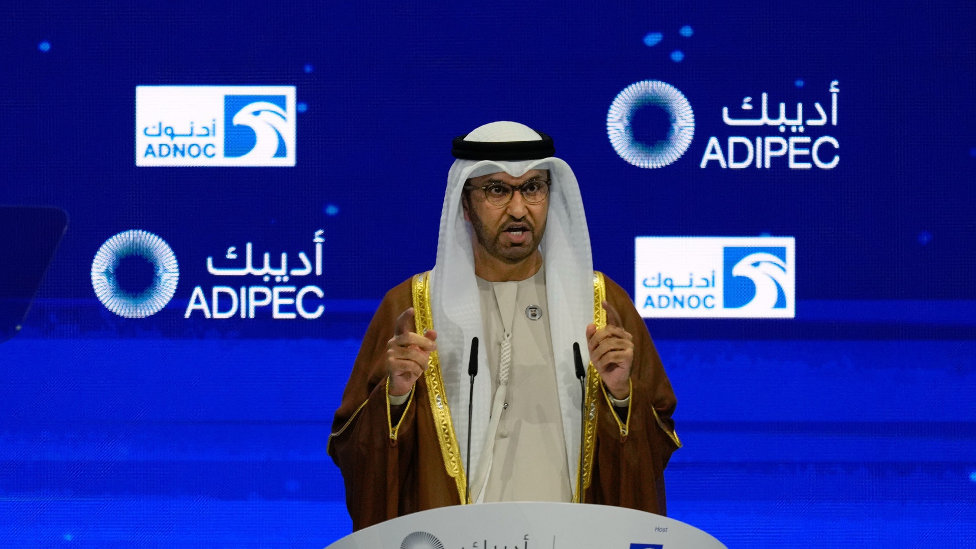 Man i traditionell arabisk klädedräkt talar vid ett podium med ADNOC och ADIPEC logotyper i bakgrunden.