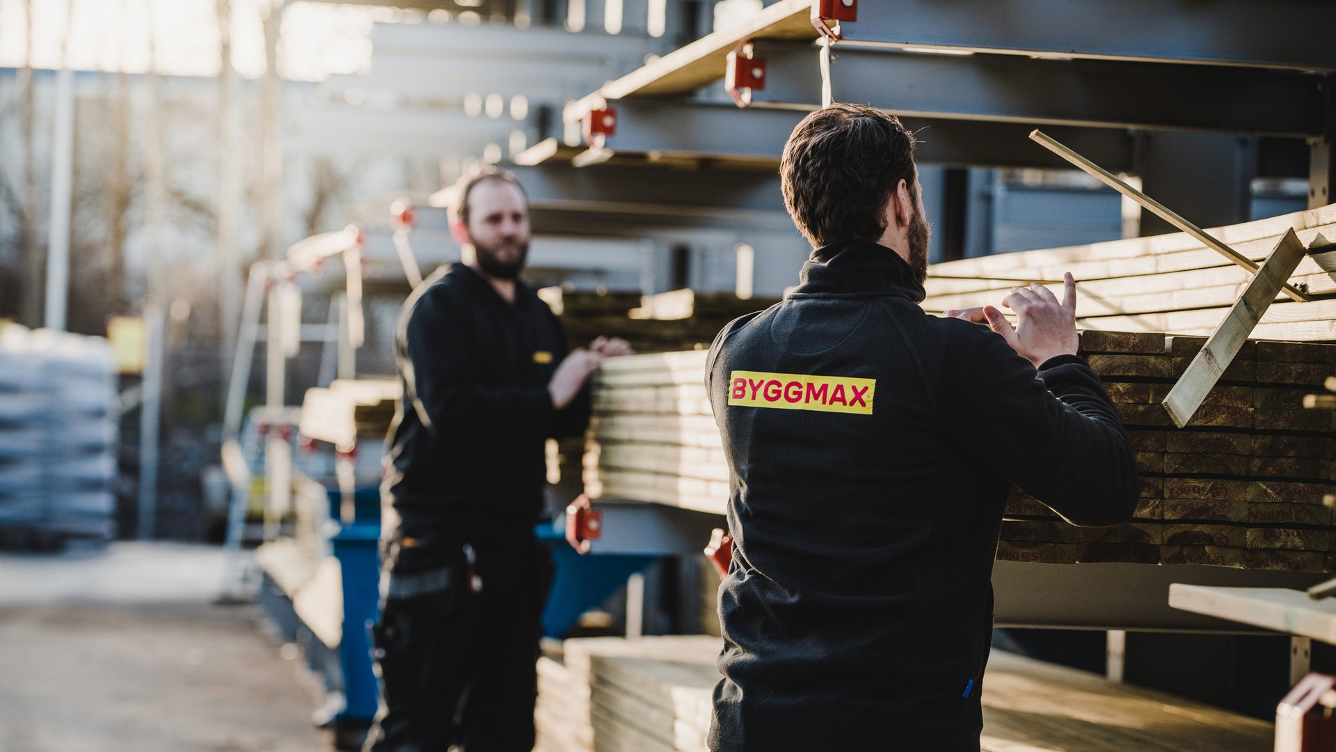 Två män i svarta arbetskläder med Byggmax-logotyp plockar bland travar av virke på ett utomhuslager.
