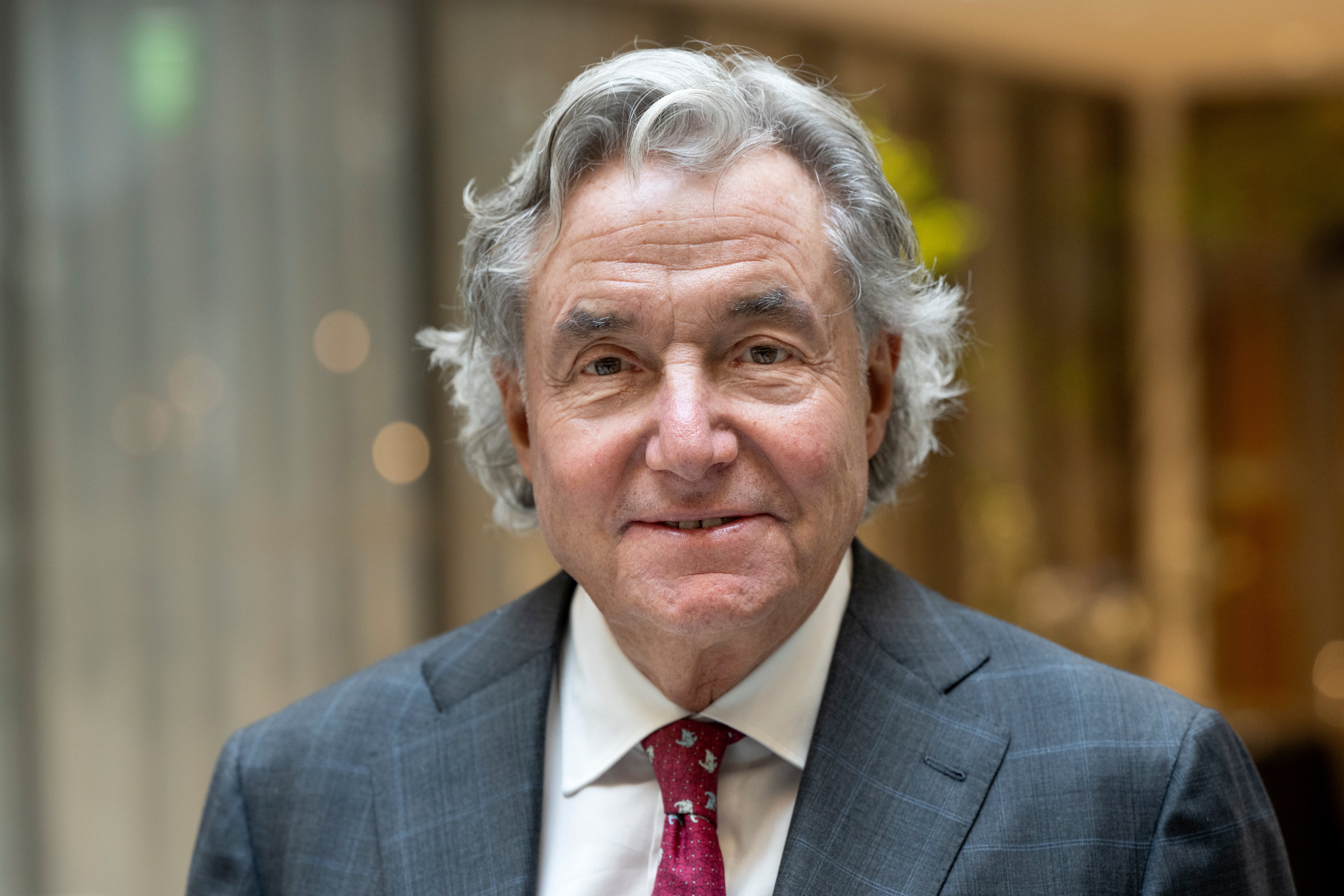 Porträtt av äldre man med grått hår, iklädd mörkblå kostym, vit skjorta och vinröd slips.