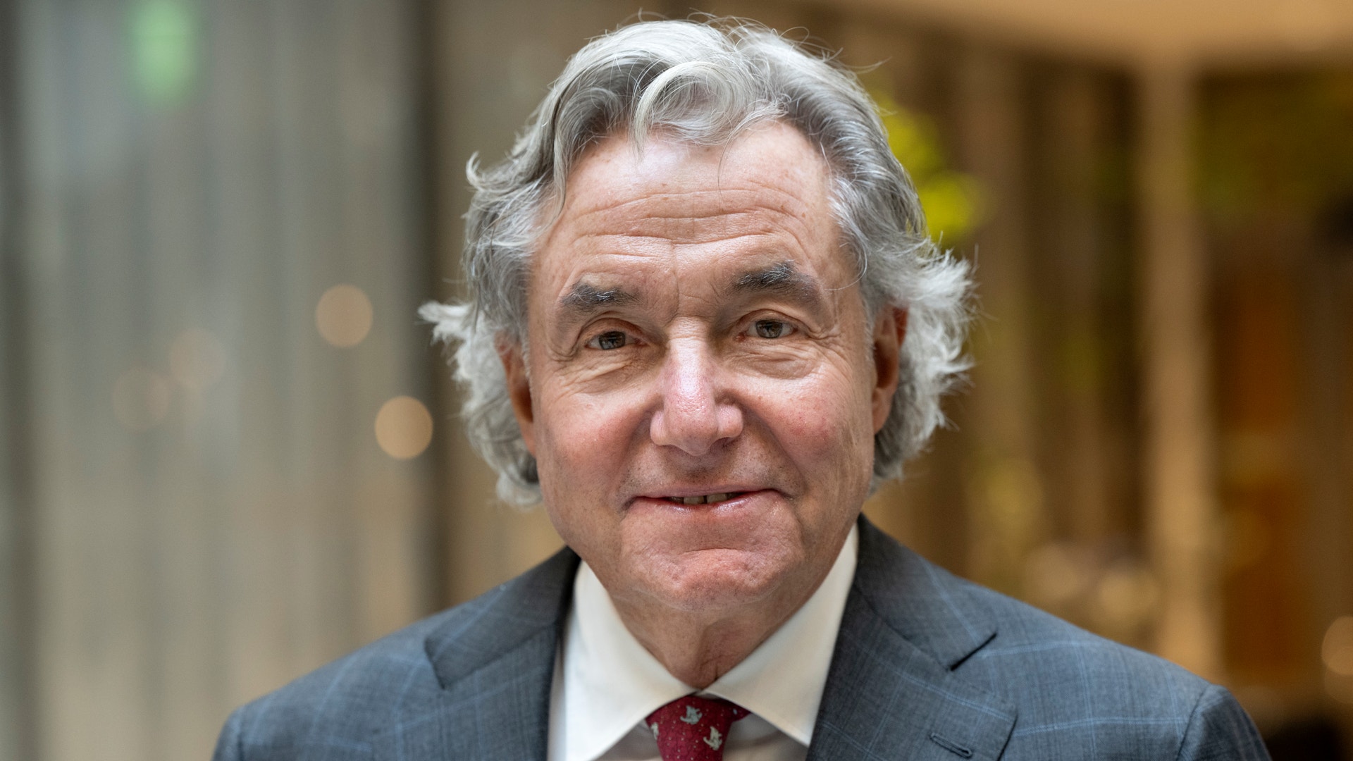 Porträtt av äldre man med grått hår, iklädd mörkblå kostym, vit skjorta och vinröd slips.