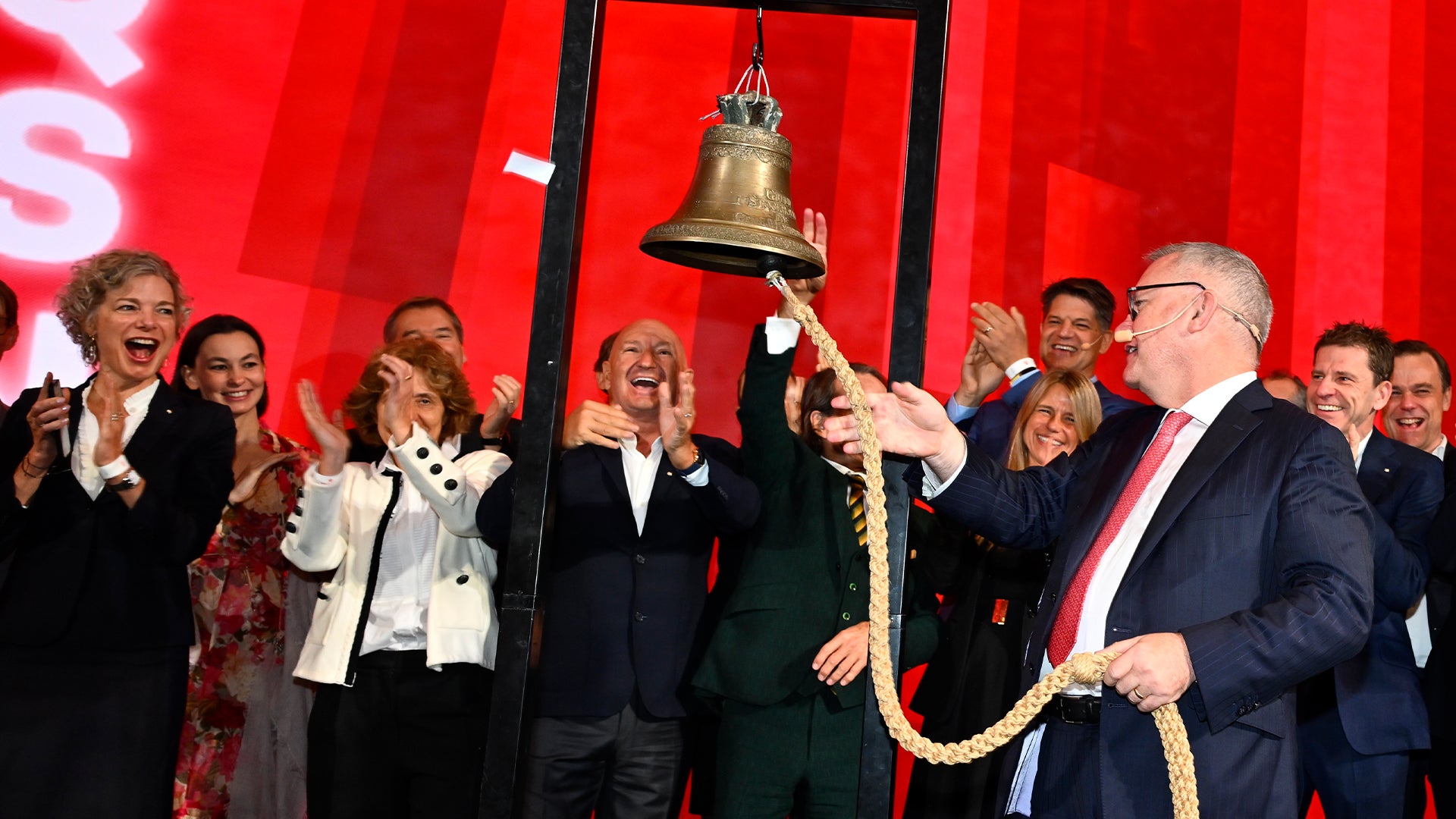 Grupp människor i formell klädsel som firar tillsammans framför röd bakgrund. En man håller i ett rep fäst vid en stor guldklocka som hänger från ovan.