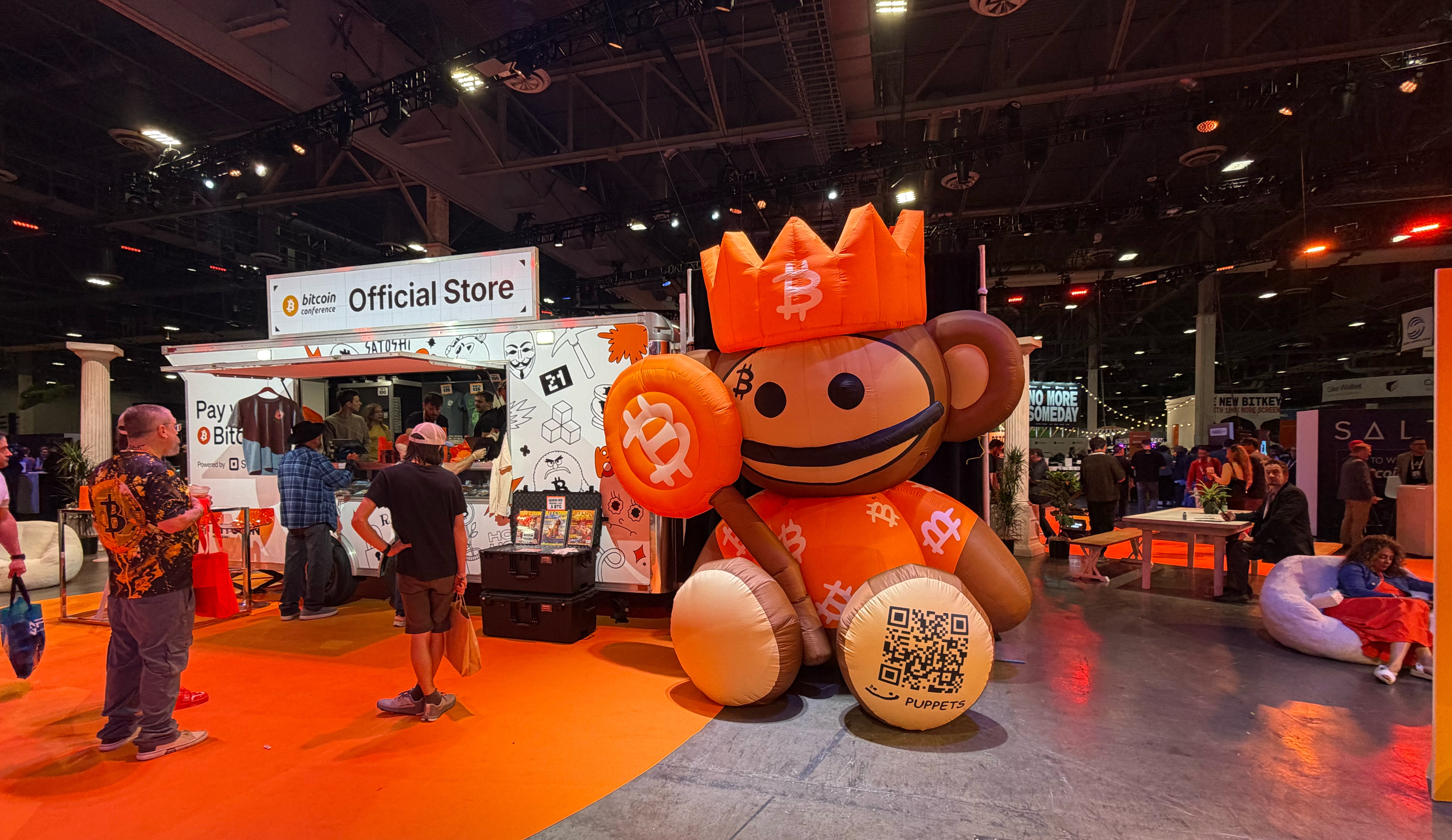 En stor orange maskot med leende ansikte och en krona på sitt huvud står på en orange mässpodium. I bakgrunden syns en officiell butik och besökare på en utställning.