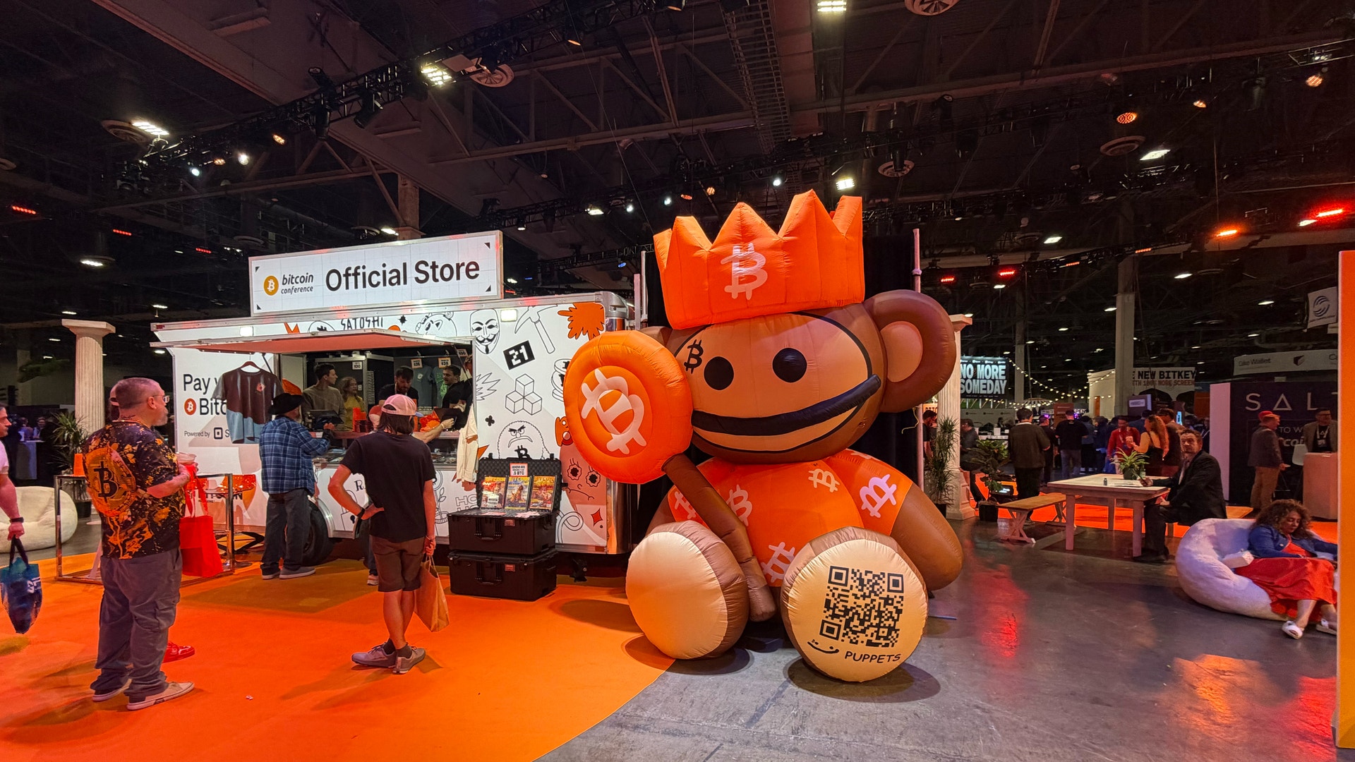 En stor orange maskot med leende ansikte och en krona på sitt huvud står på en orange mässpodium. I bakgrunden syns en officiell butik och besökare på en utställning.