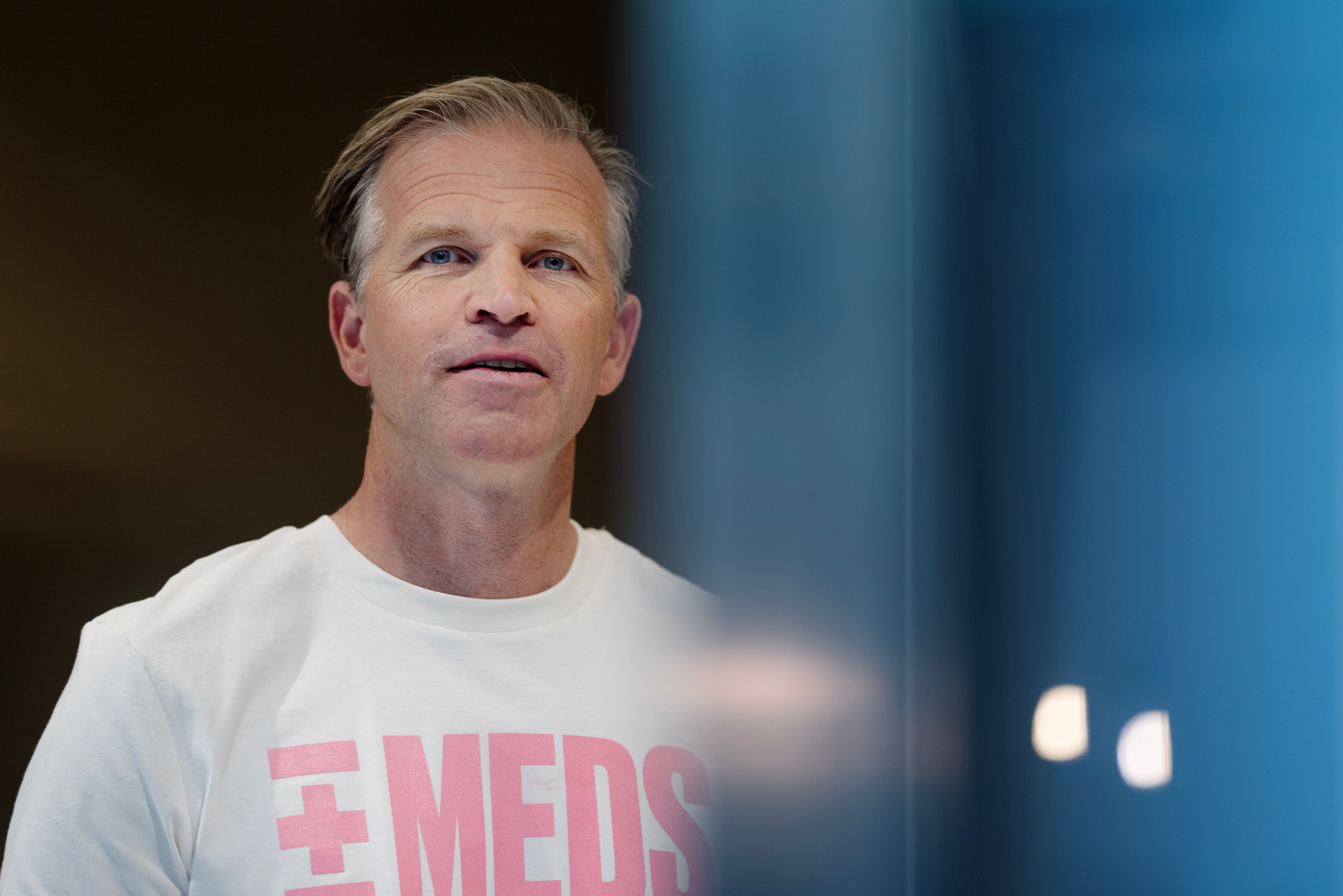 Porträtt av en man i vit t-shirt med rosa text, fotograferad mot en suddig bakgrund.