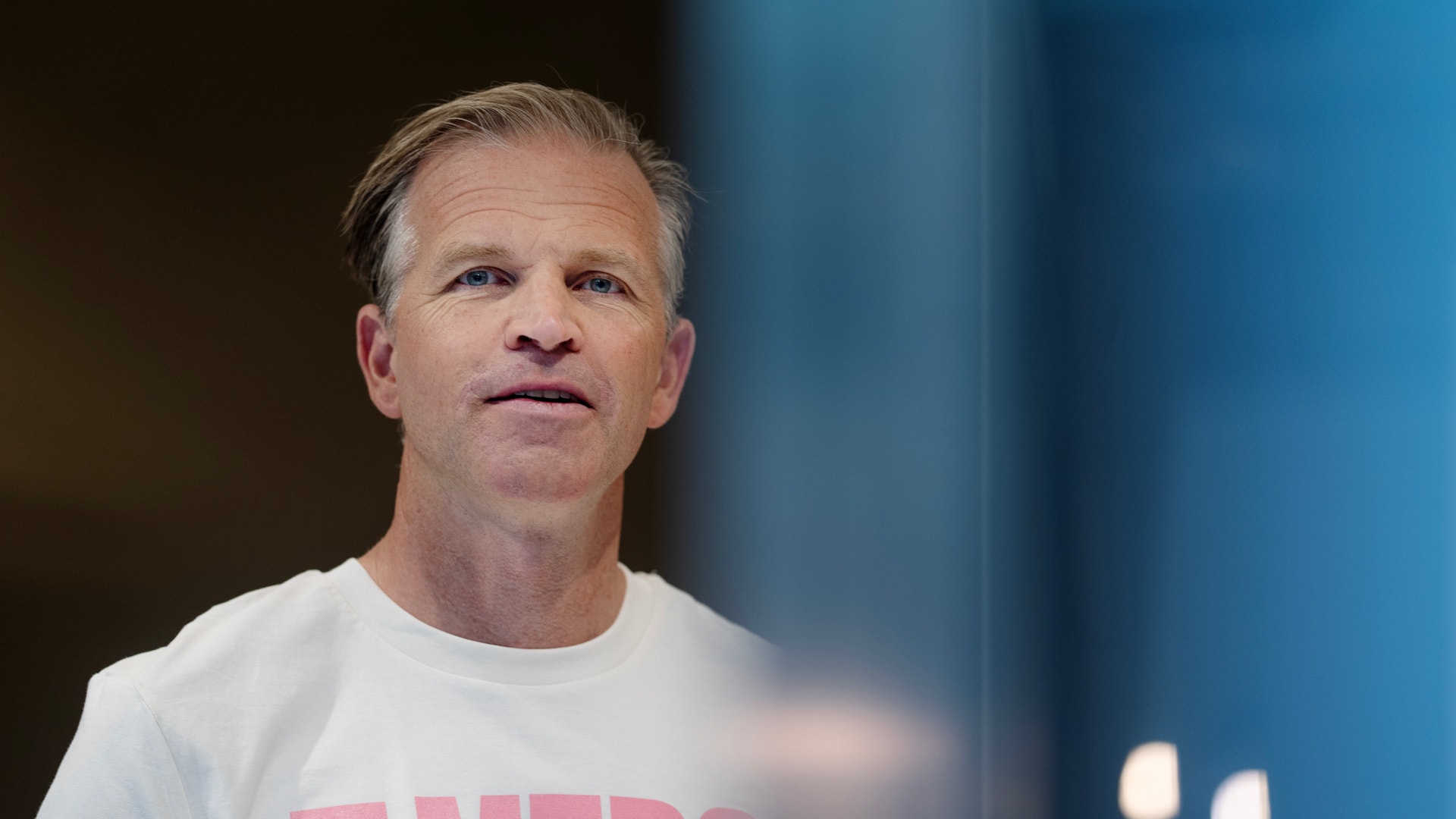 Porträtt av en man i vit t-shirt med rosa text, fotograferad mot en suddig bakgrund.