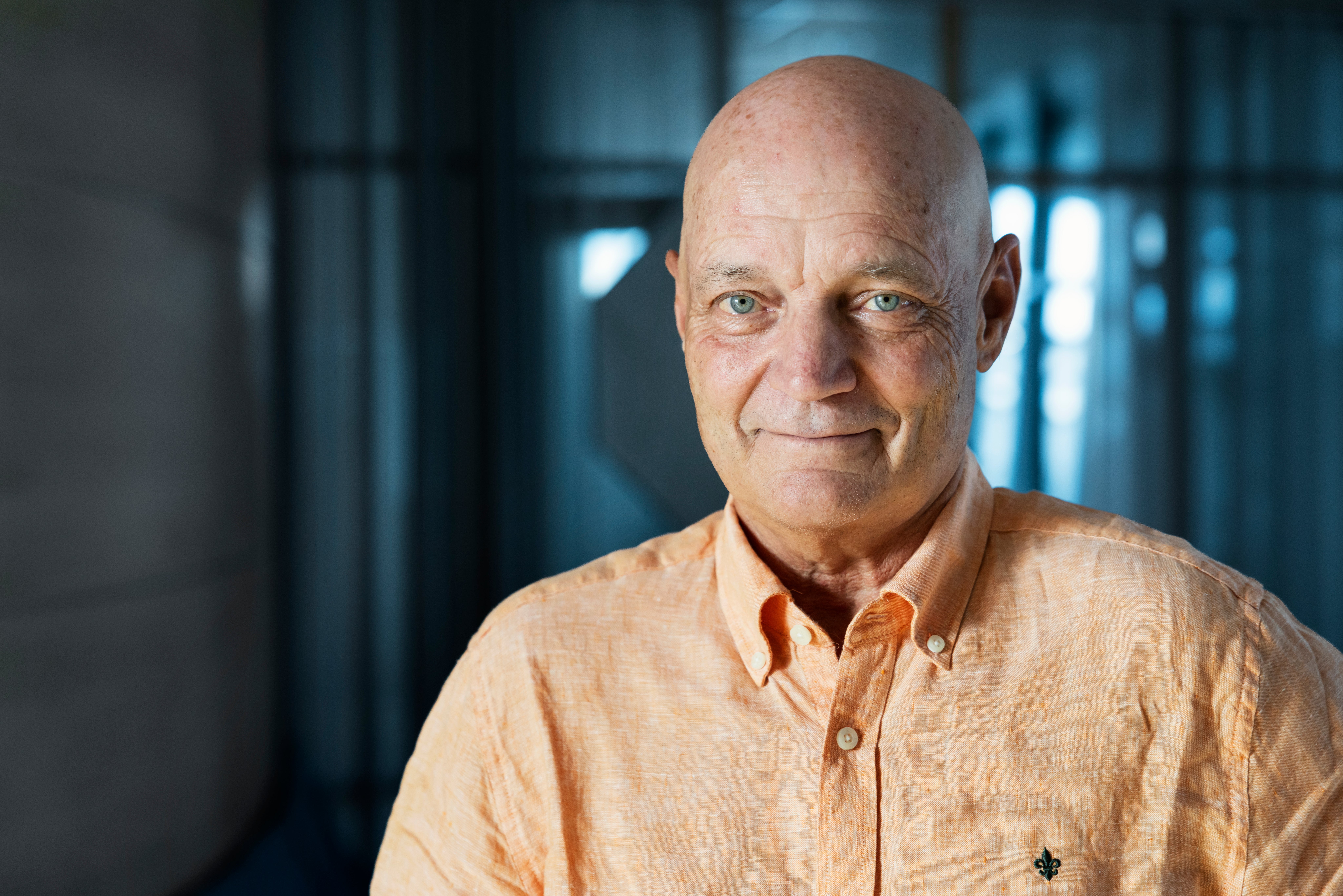 Porträtt av en man som ler mot kameran. Han bär en ljus orange skjorta och fotograferas  inomhus.