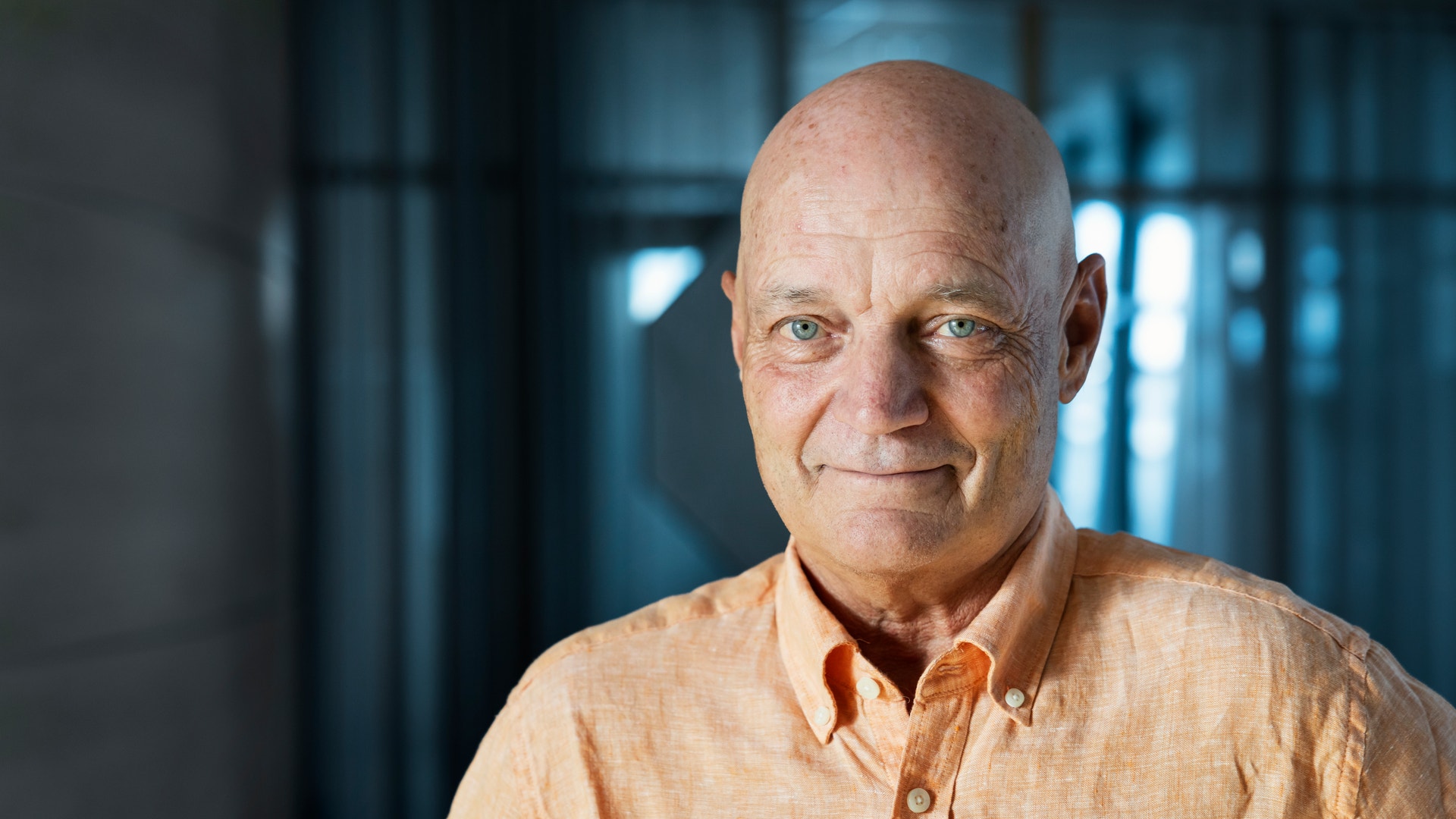 Porträtt av en man som ler mot kameran. Han bär en ljus orange skjorta och fotograferas inomhus.
