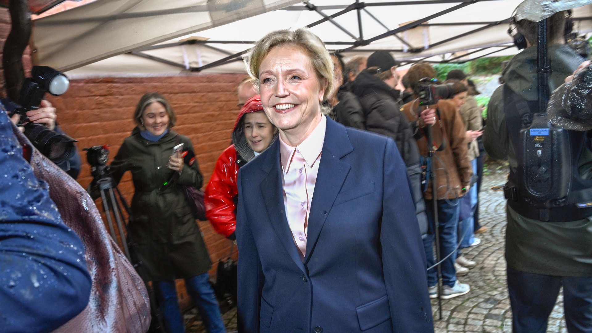 Elisabeth Thand Ringqvist, partiledare för Centerpartiet, ler när hon går utomhus i regn, omgiven av fotografer och journalister.