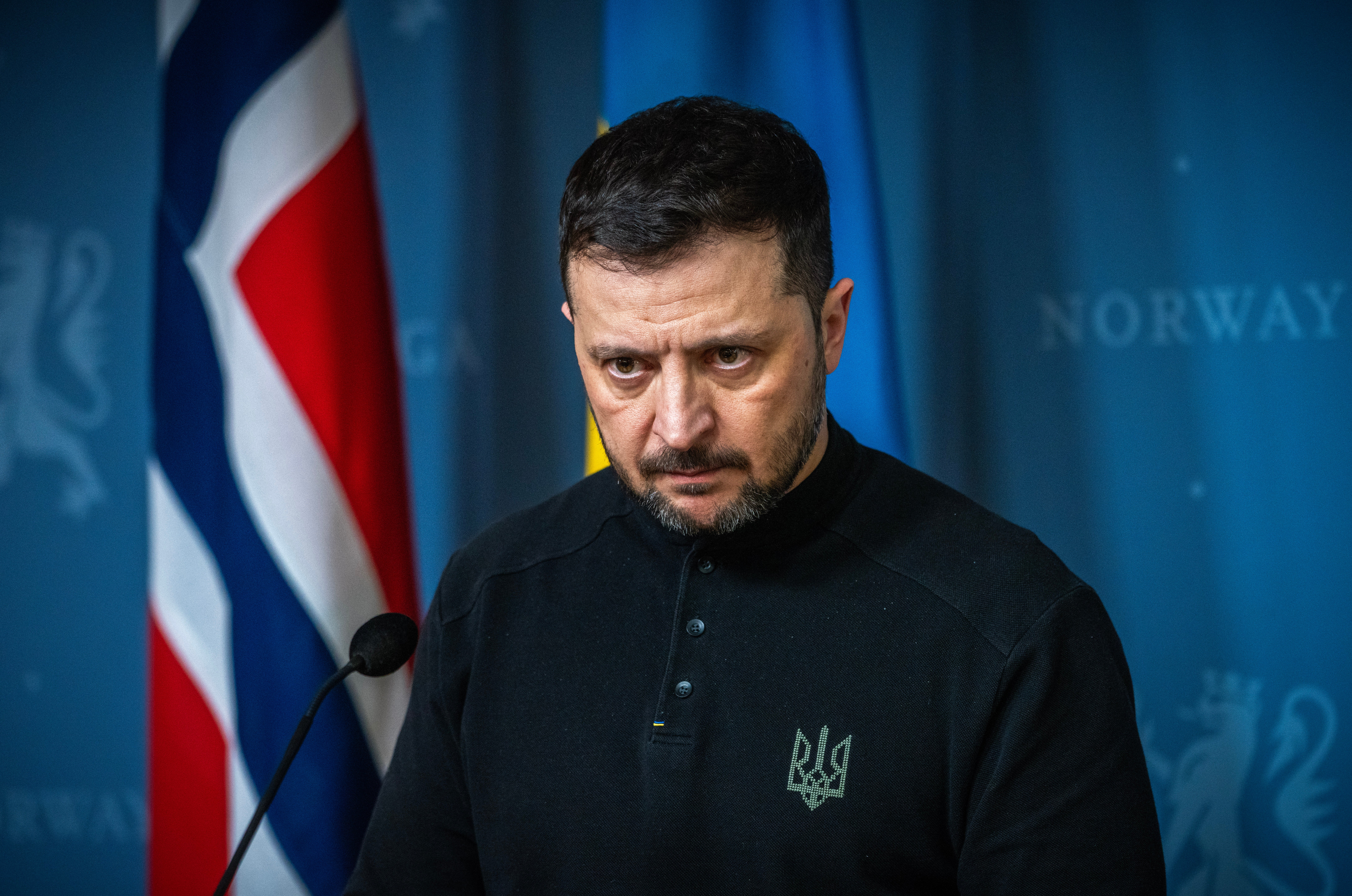 Ukrainas president Volodymyr Zelenskyj står allvarlig vid en mikrofon framför en norsk och en ukrainsk flagga.