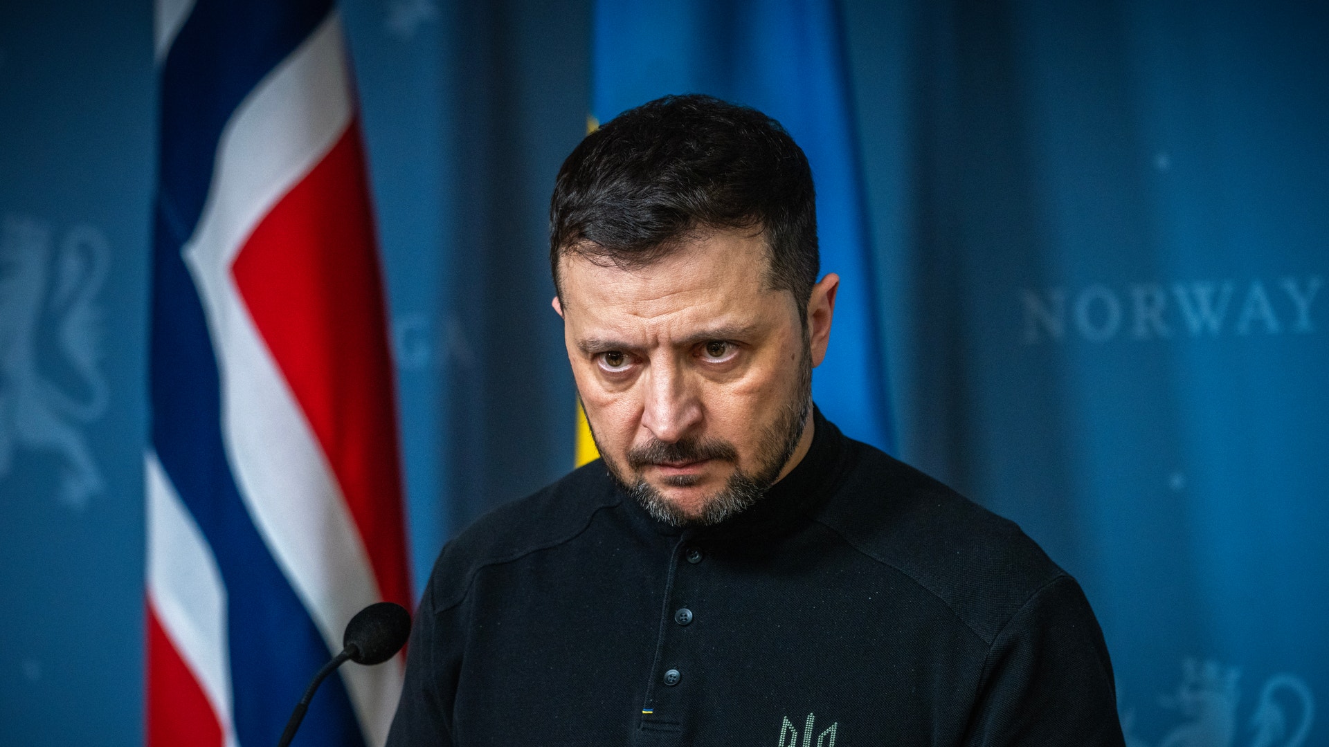 Ukrainas president Volodymyr Zelenskyj står allvarlig vid en mikrofon framför en norsk och en ukrainsk flagga.