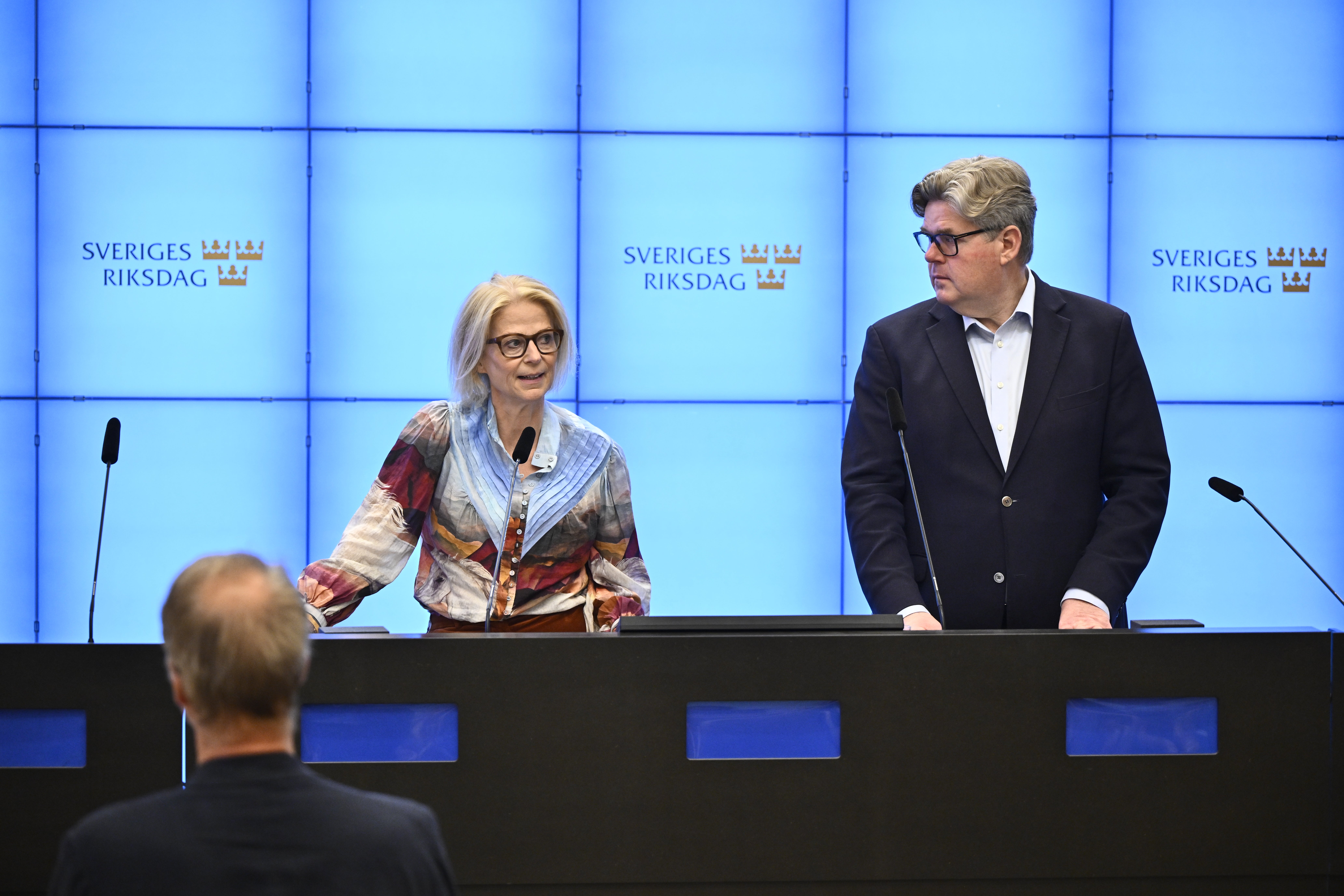 Två personer vid ett podium under en presskonferens på Sveriges riksdag. En person i färgglad tröja och glasögon står till vänster, en man i mörk kostym står till höger. Blå vägg med riksdagslogotyper i bakgrunden.