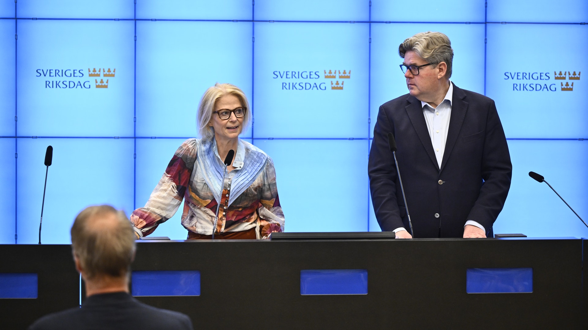 Två personer vid ett podium under en presskonferens på Sveriges riksdag. En person i färgglad tröja och glasögon står till vänster, en man i mörk kostym står till höger. Blå vägg med riksdagslogotyper i bakgrunden.