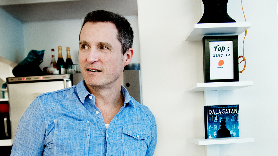 Porträtt av en man i ljusblå skjorta som står framför en vägg med vita hyllor innehållande böcker och dekorativa föremål. Mannen tittar mot kameran med ett vänligt uttryck.