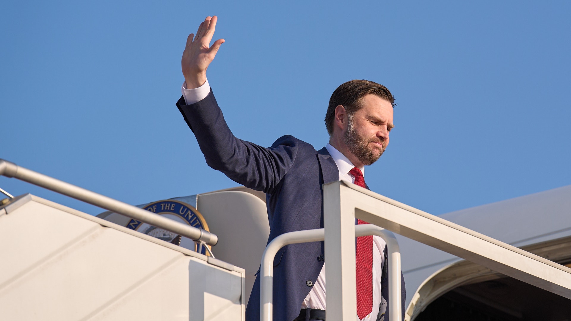 Man i mörk kostym och röd slips vinkar från flygplanets trappa mot blå himmel.