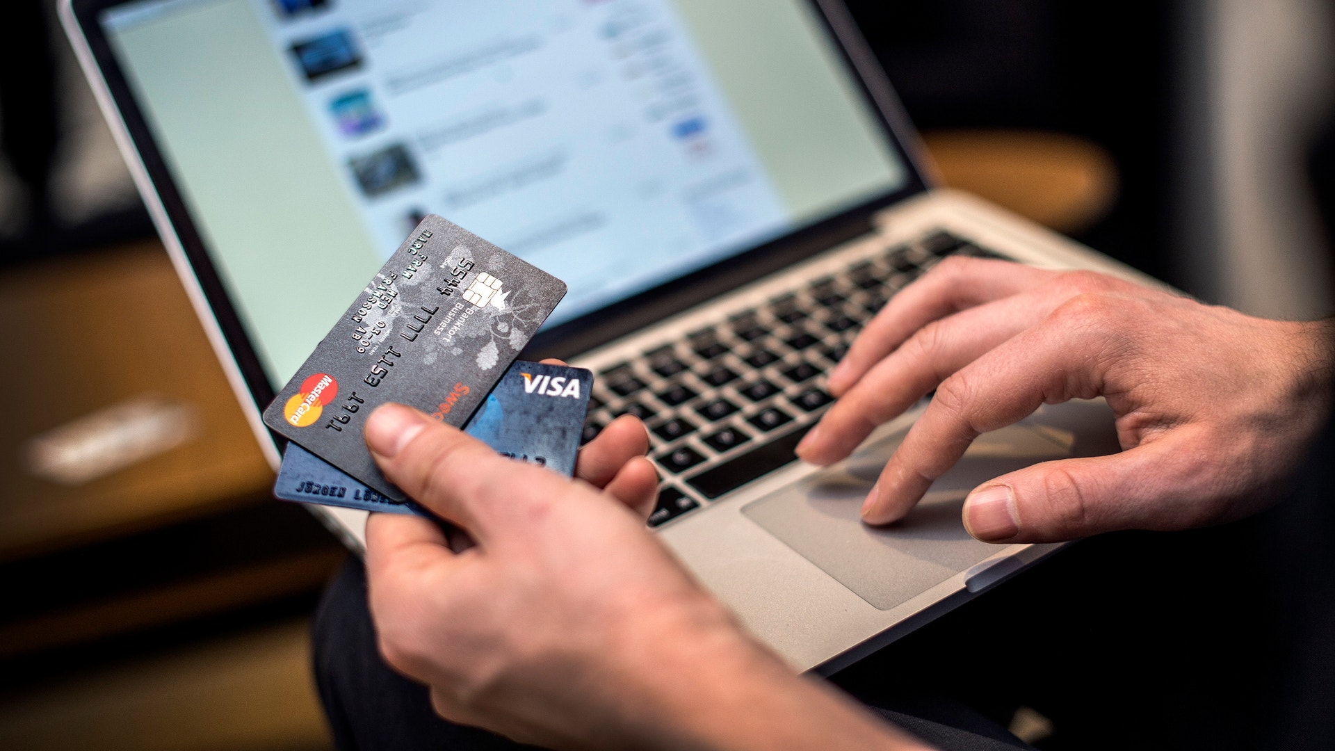 En person håller ett kreditkort framför en laptop-skärm som visar en webbutik eller betalningssida. Personens högerhand vilar på tangentbordet medan vänsterhand håller kortet. Bilden illustrerar online-shopping, e-handel och säker digital betalning.