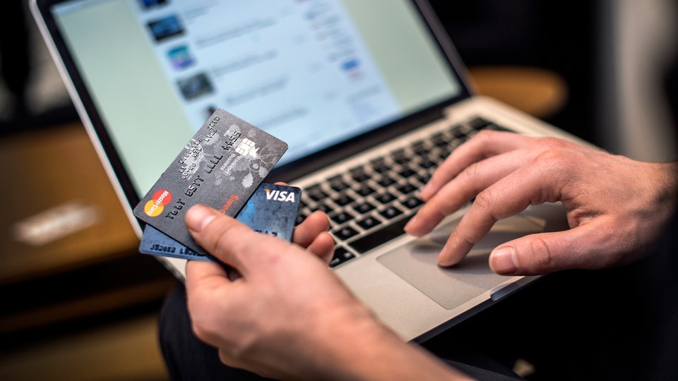 En person håller ett kreditkort framför en laptop-skärm som visar en webbutik eller betalningssida. Personens högerhand vilar på tangentbordet medan vänsterhand håller kortet. Bilden illustrerar online-shopping, e-handel och säker digital betalning.