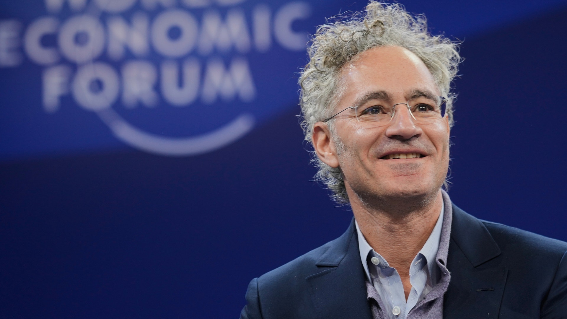 Man med grått lockigt hår och glasögon i mörk kavaj framför World Economic Forum-bakgrund.