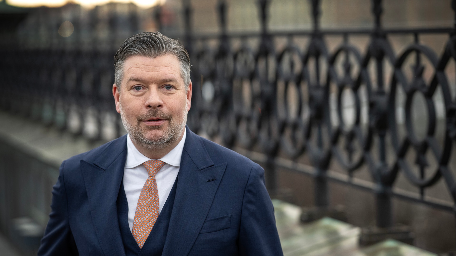 Porträtt av en man i mörkblå kostym och slips som står utomhus framför ett svart smidesräcke och tittar in i kameran.
