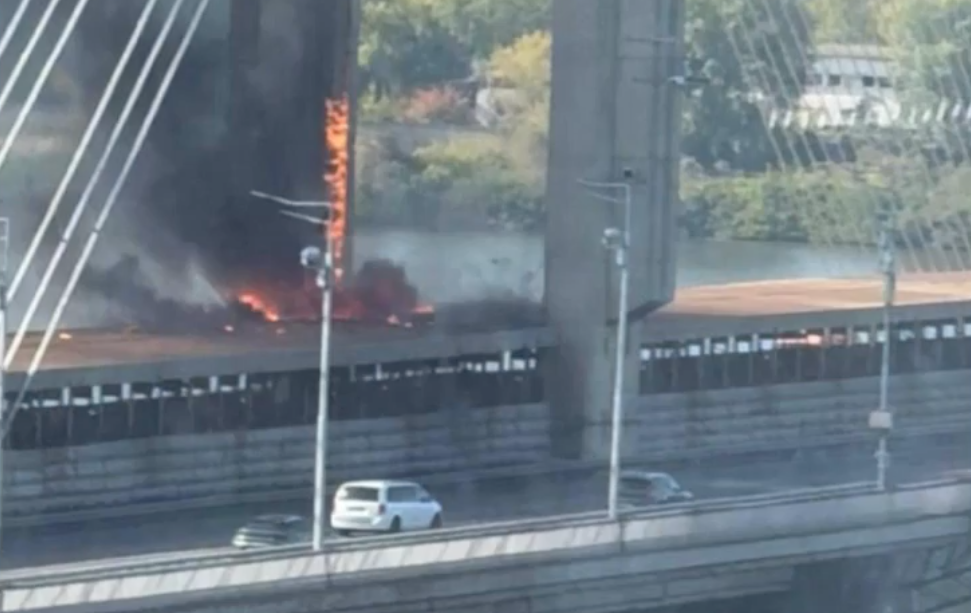 Snedkabelbro med stora betongpyloner där en kraftig brand med lågor och tjock rök syns på brobanan, medan bilar kör förbi.