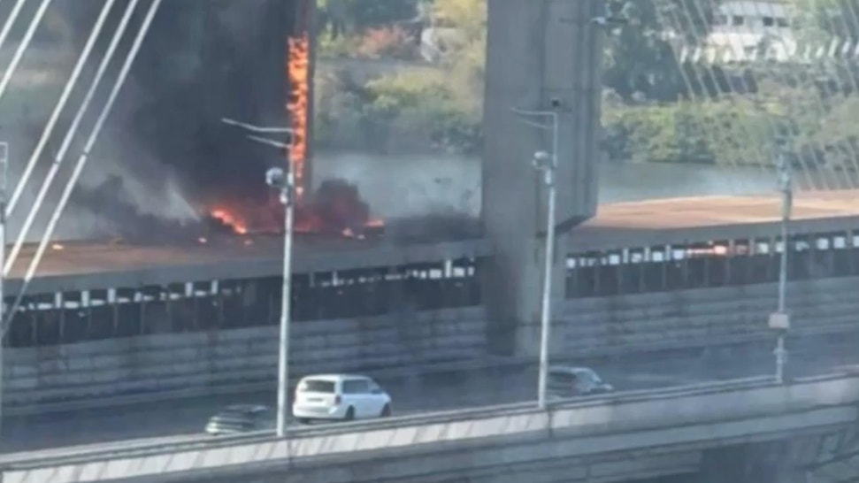 Snedkabelbro med stora betongpyloner där en kraftig brand med lågor och tjock rök syns på brobanan, medan bilar kör förbi.