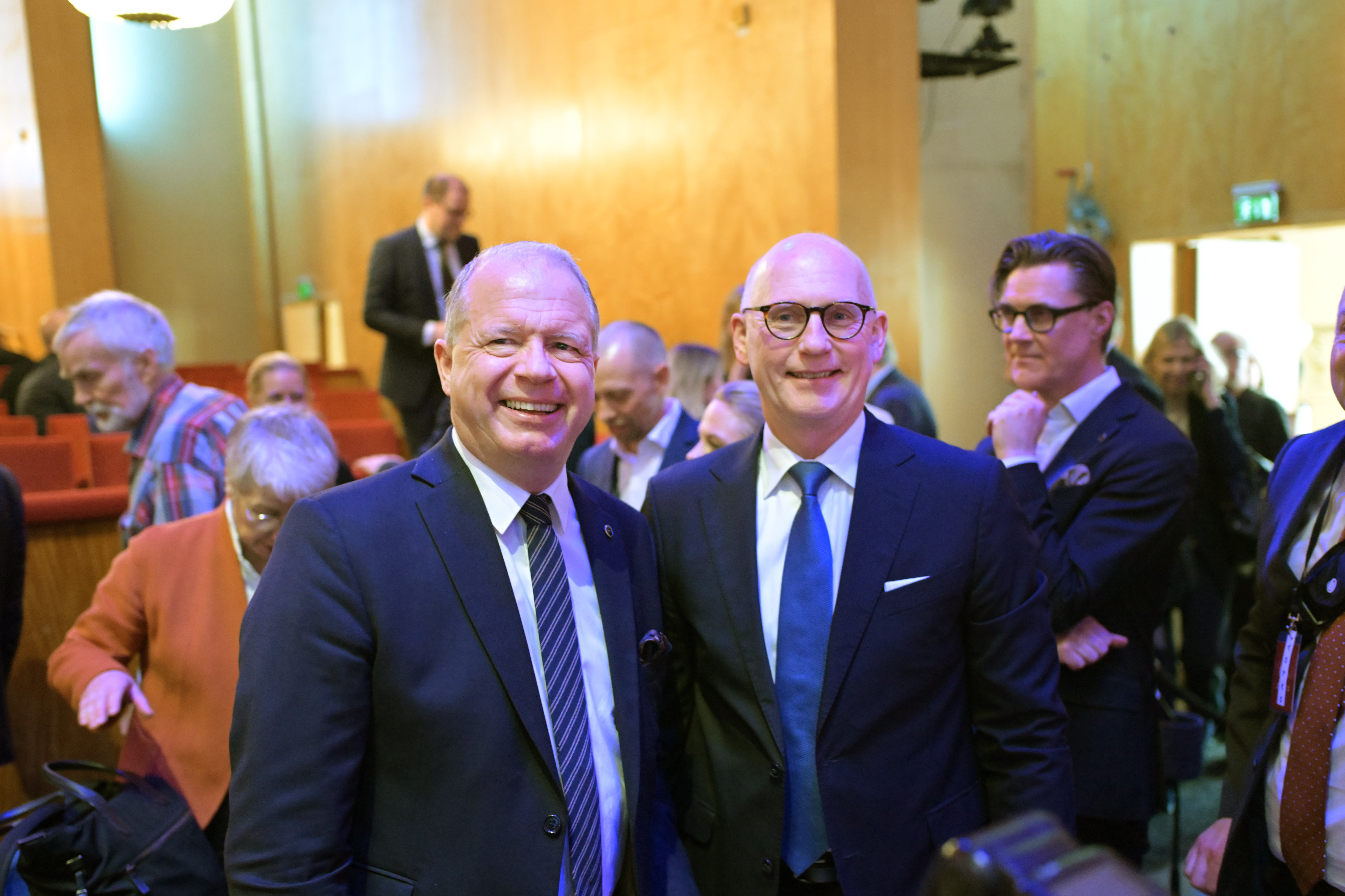 Två män i kostym och slips står framför kameran och ler på ett formellt event. I bakgrunden syns fler människor i en konferenssal.
