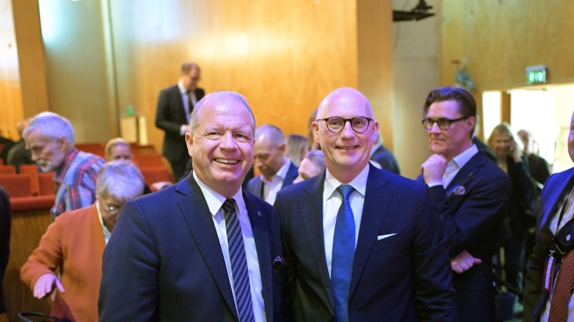 Två män i kostym och slips står framför kameran och ler på ett formellt event. I bakgrunden syns fler människor i en konferenssal.