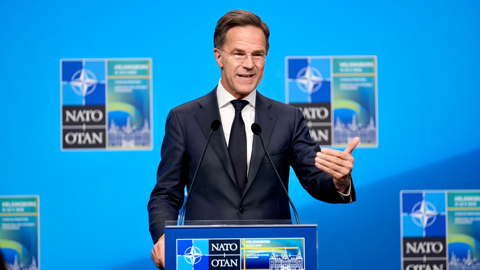 Man i mörk kostym och slips står vid NATO-podium och håller tal. Blå bakgrund med NATO-loggor syns bakom honom.