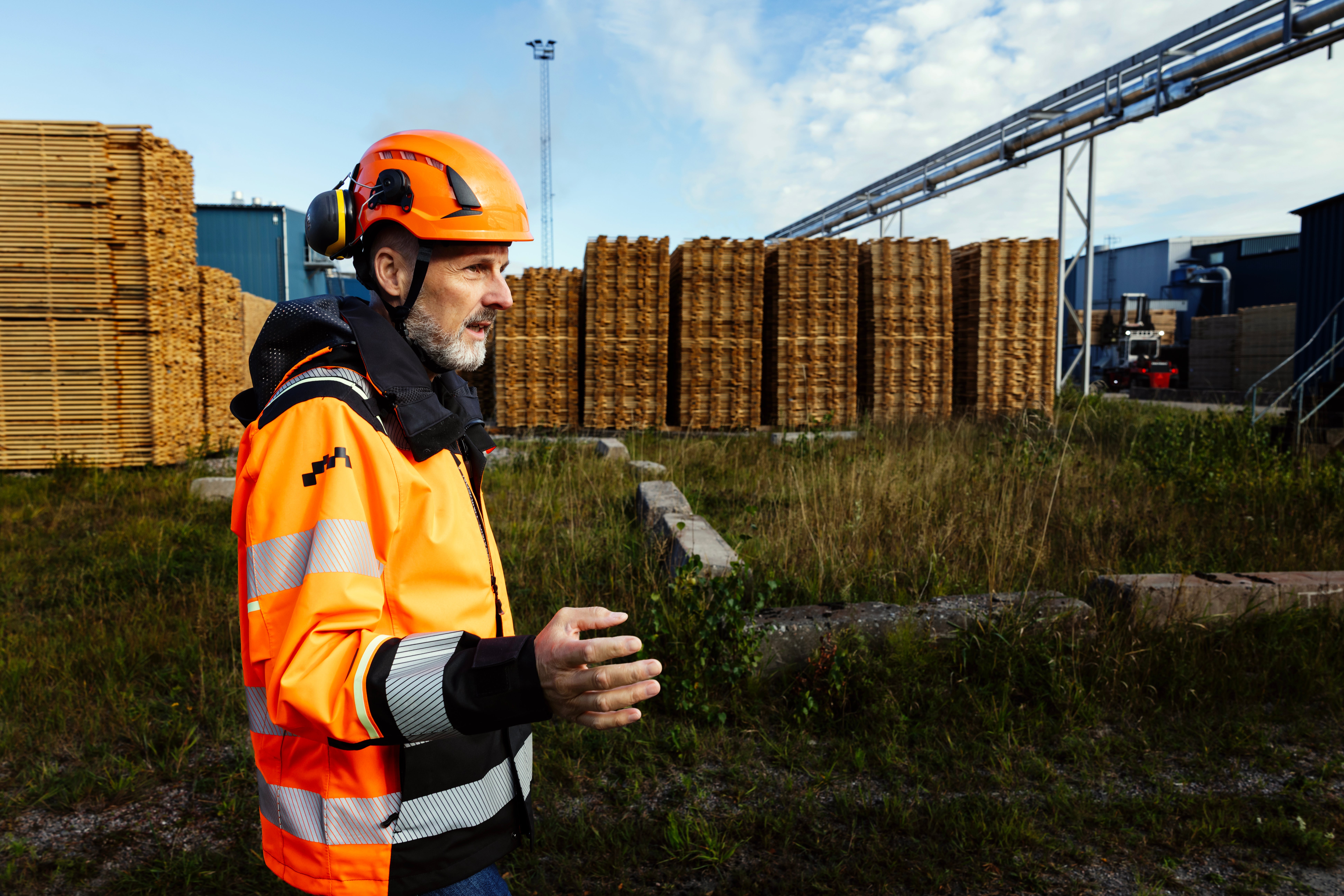 En man i orange skyddskläder och hjälm står vid ett sågverk med staplade timmerpaket i bakgrunden.